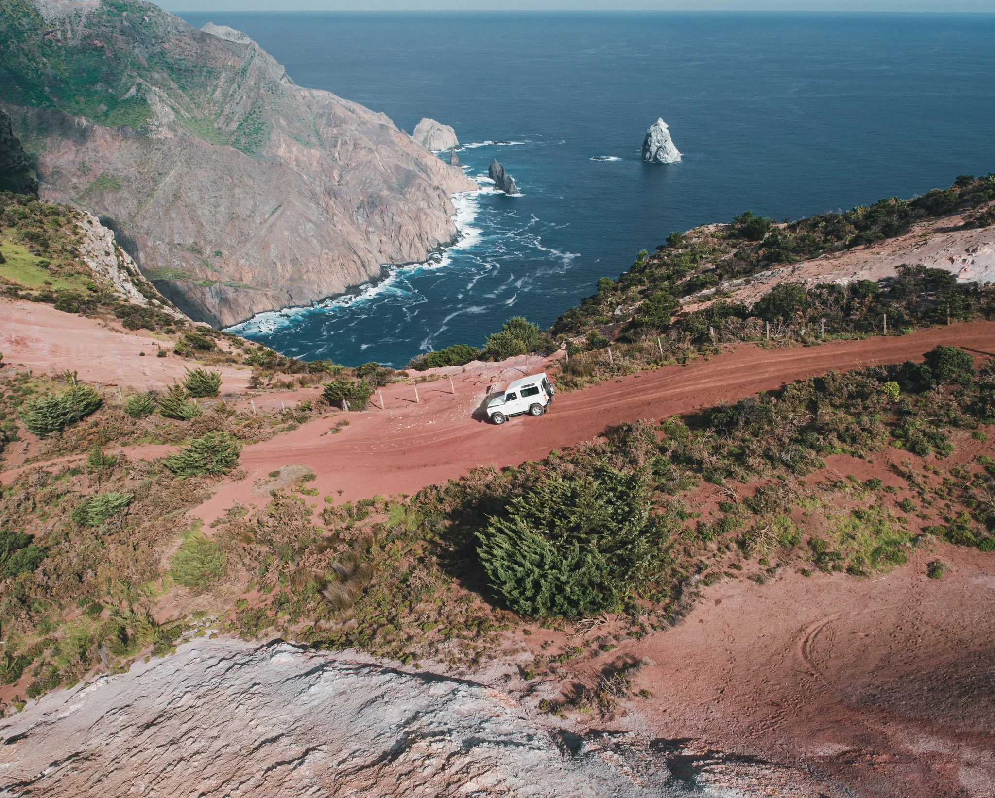 A white vehicle driving on a dirt road on a hillside overlooking the ocean with rocky cliffs and distant rocks in the water.