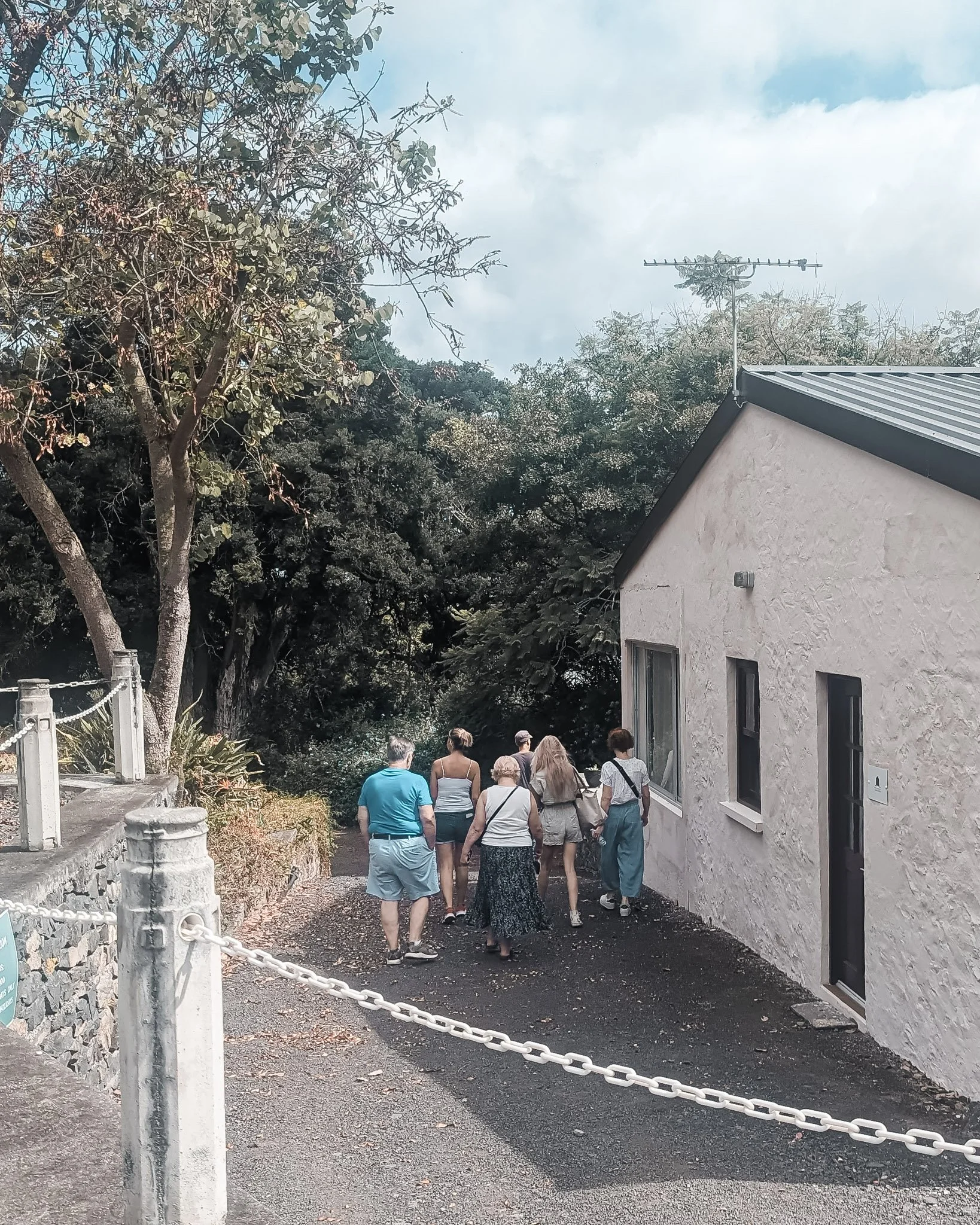 A group of seven people walking along a gravel path past a white stucco house with black window frames, surrounded by trees and greenery.