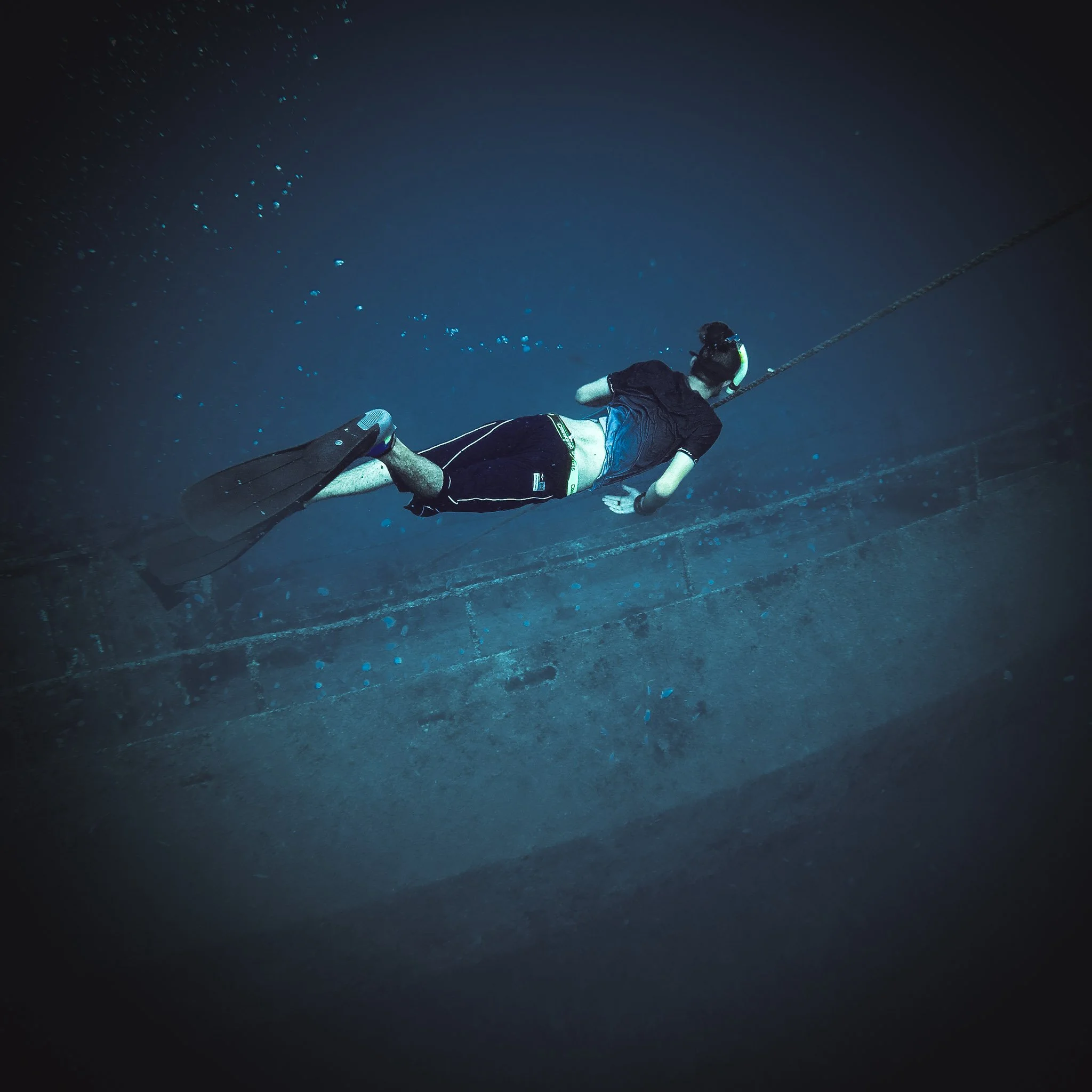 Person scuba diving in a blue underwater environment, wearing fins, a wetsuit, and a diving mask, holding onto a line attached to the surface.