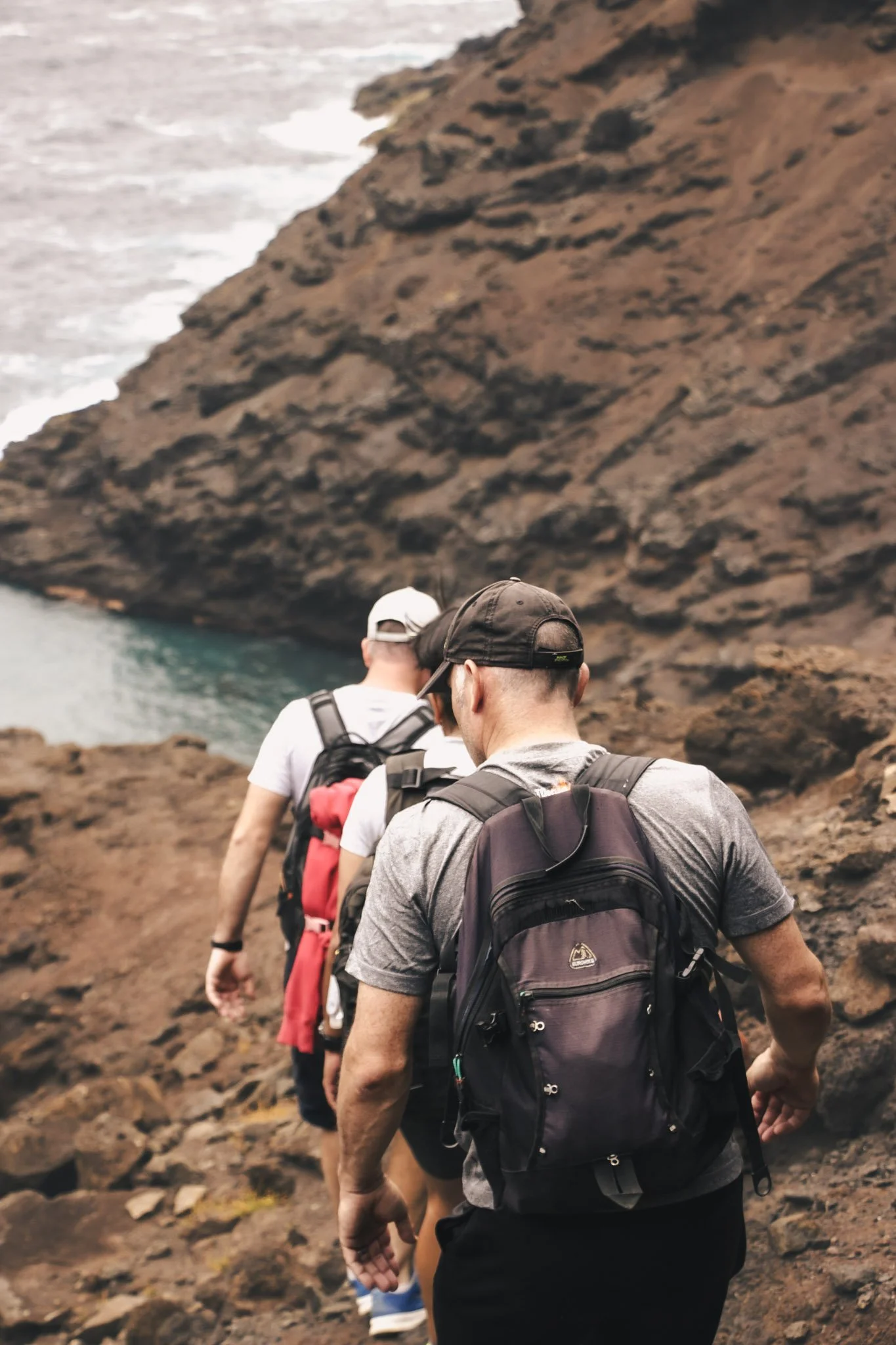Three hikers walking on a rocky trail along a coastline, with a large cliff on one side and the ocean in the background.
