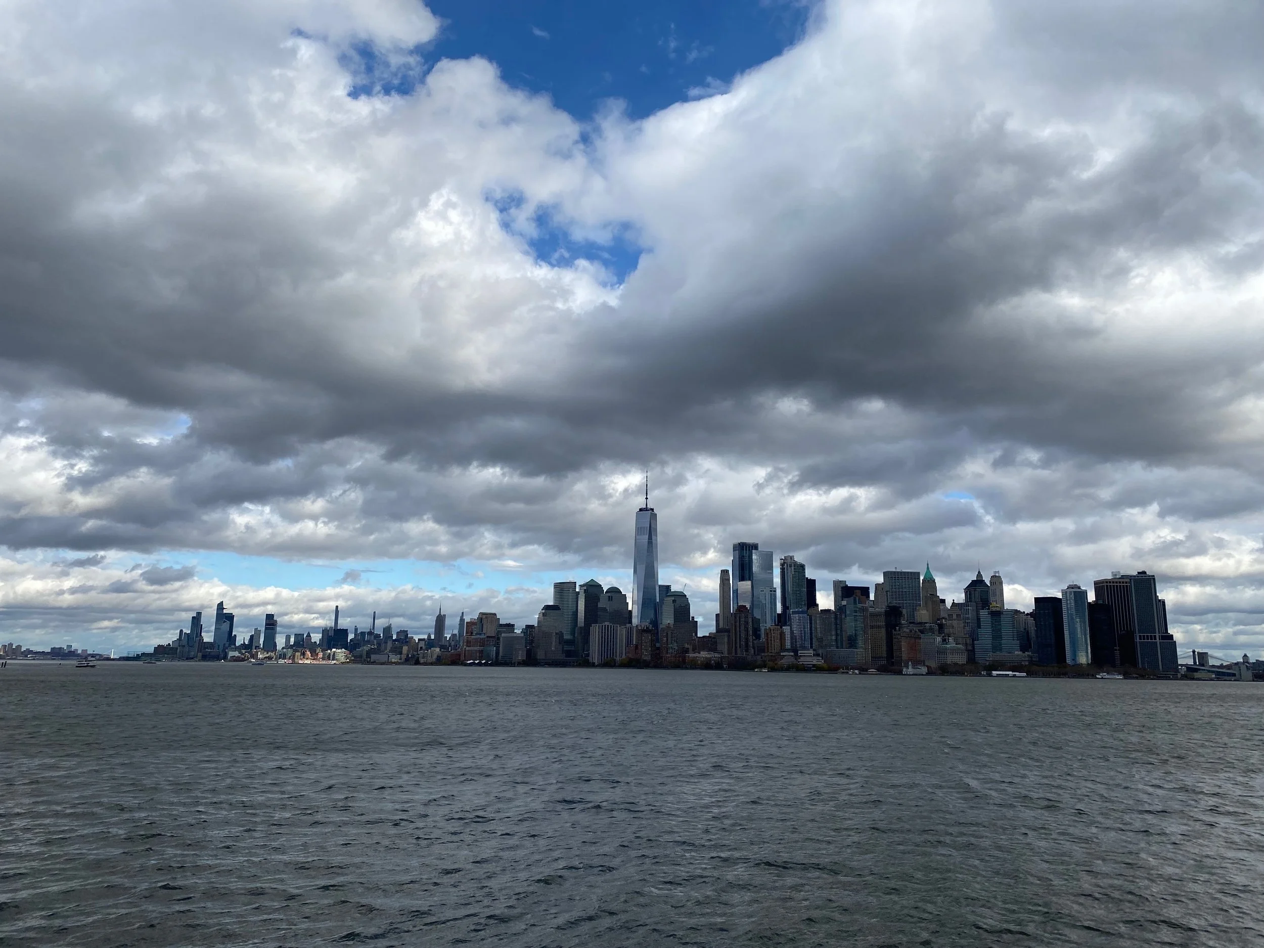 Skyline of New York City with tall buildings under a cloudy sky, viewed across the water.