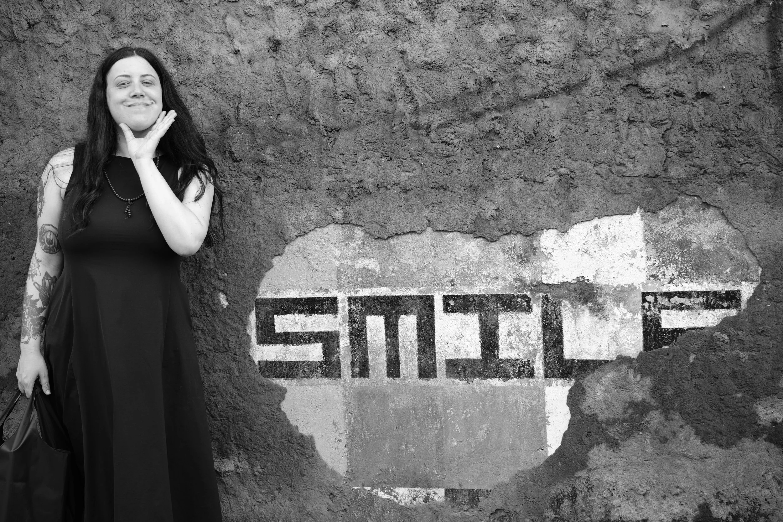 A woman with long dark hair and tattoos on her right arm stands against a textured concrete wall with a partially visible painted sign. She is smiling subtly and resting her left hand on her face, wearing a sleeveless black dress and a necklace.