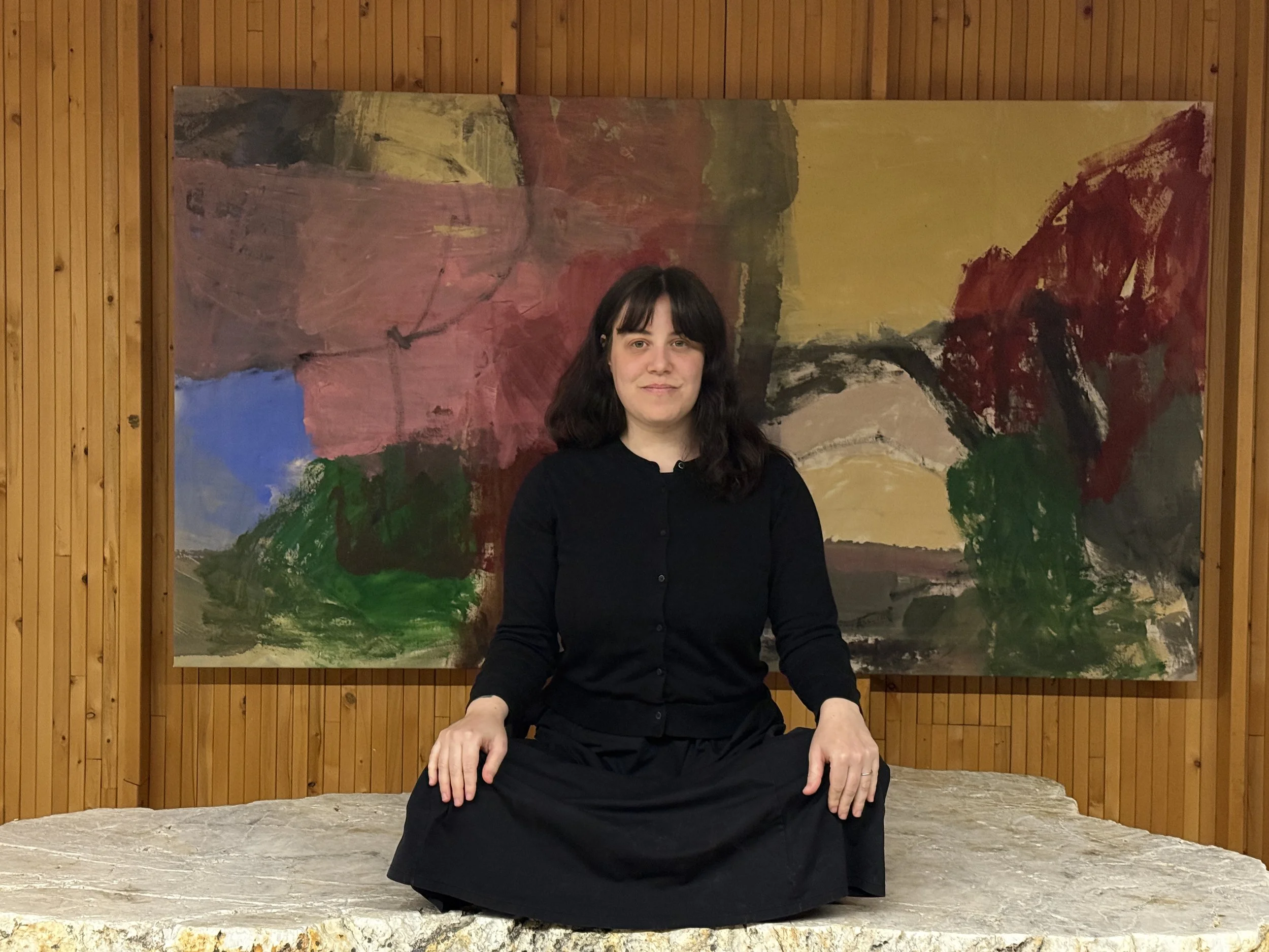 A woman with dark hair and bangs, dressed in black, sitting cross-legged in front of an abstract painting with bold colors including yellow, red, green, blue, and black, on a wooden-paneled wall.