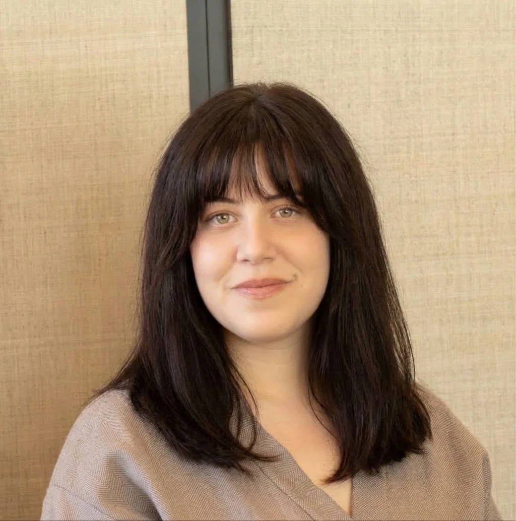 A woman with dark brown, shoulder-length hair and bangs, smiling softly, standing in a room with beige walls.