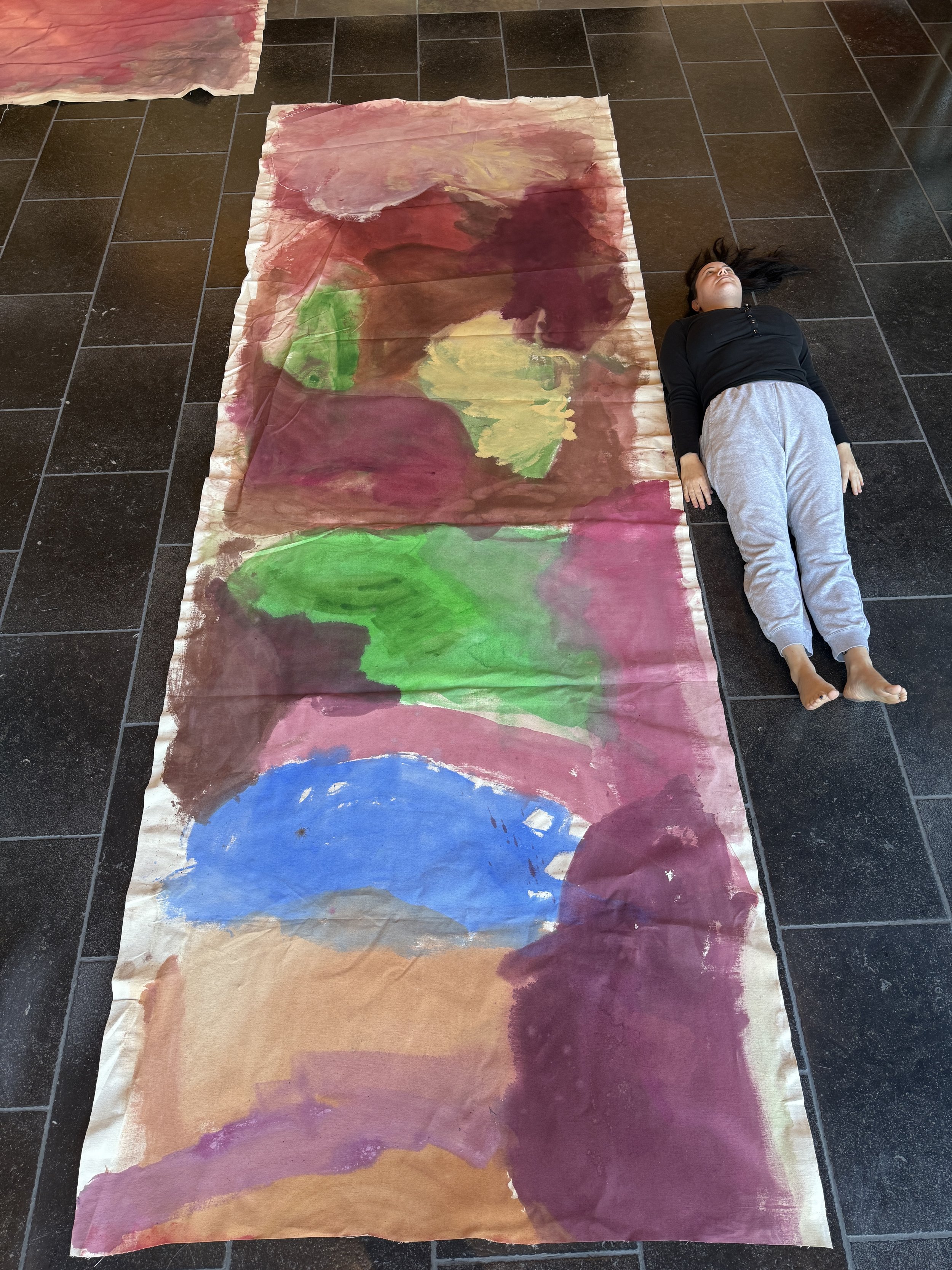 A large abstract watercolor painting laid out on the floor, with a girl lying beside it. The watercolor features various colors including red, green, yellow, blue, purple, and brown, with soft blended shapes and brushstrokes.