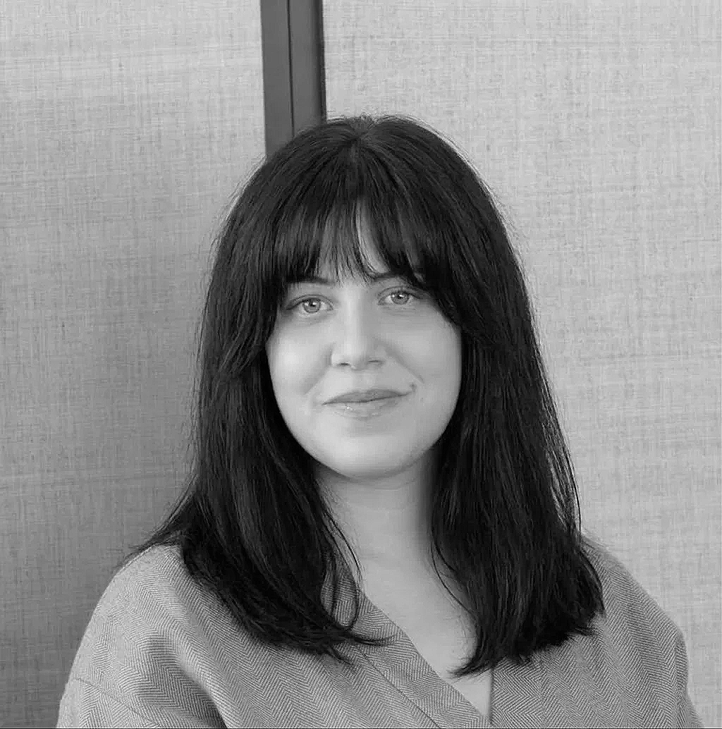 Black and white portrait of a woman with long dark hair and bangs, smiling softly, wearing a light-colored top, standing against a textured background.