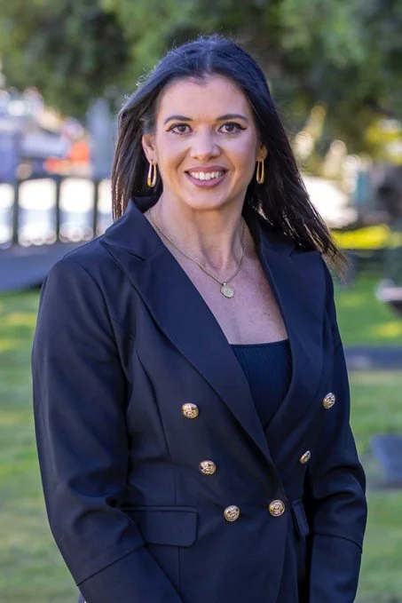 A woman with long dark hair wearing a navy blazer with gold buttons, gold hoop earrings, and a gold pendant necklace, standing outdoors with blurred trees and park scenery in the background.