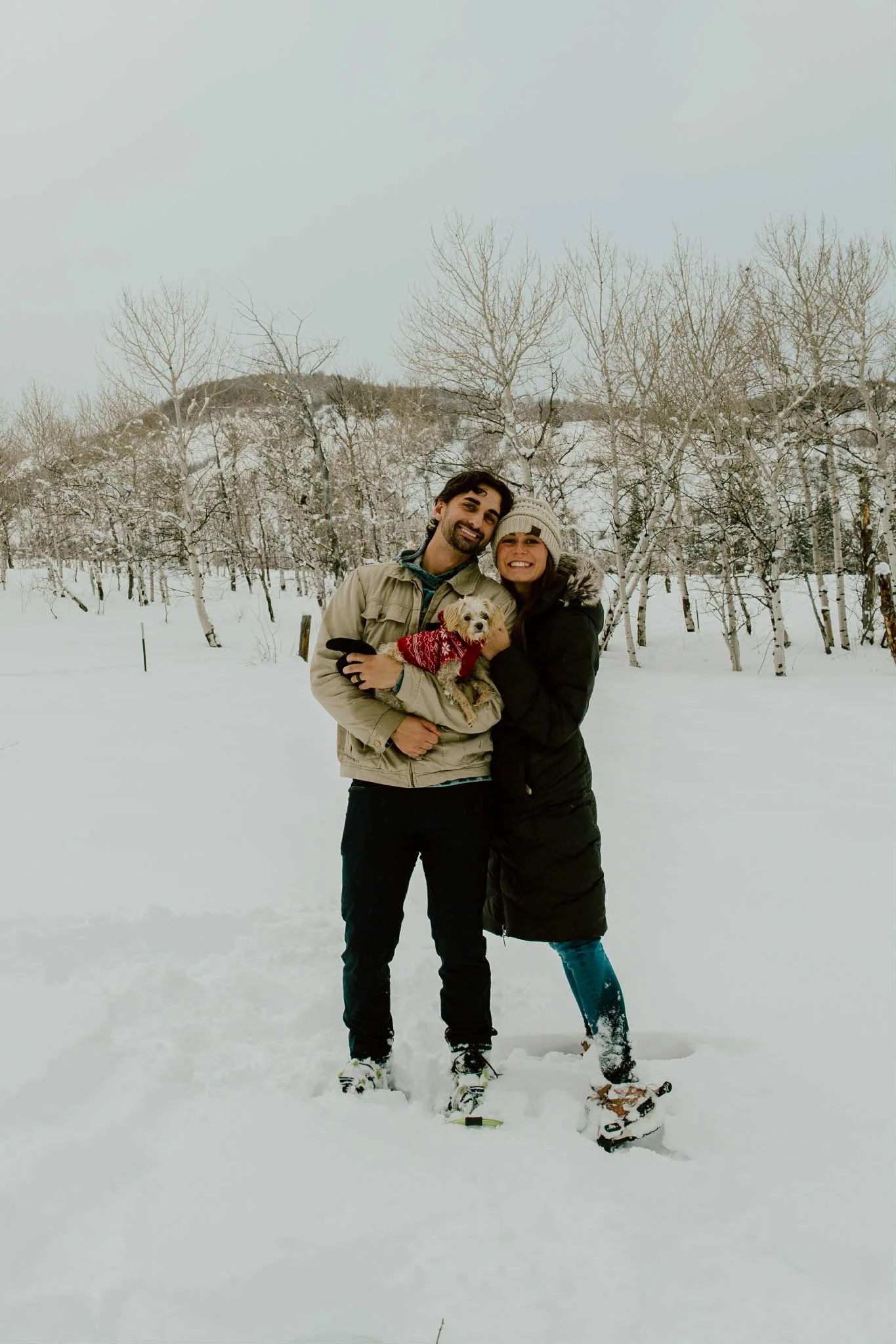 A smiling couple standing in the snow with a small dog, in a wintery woodland landscape.