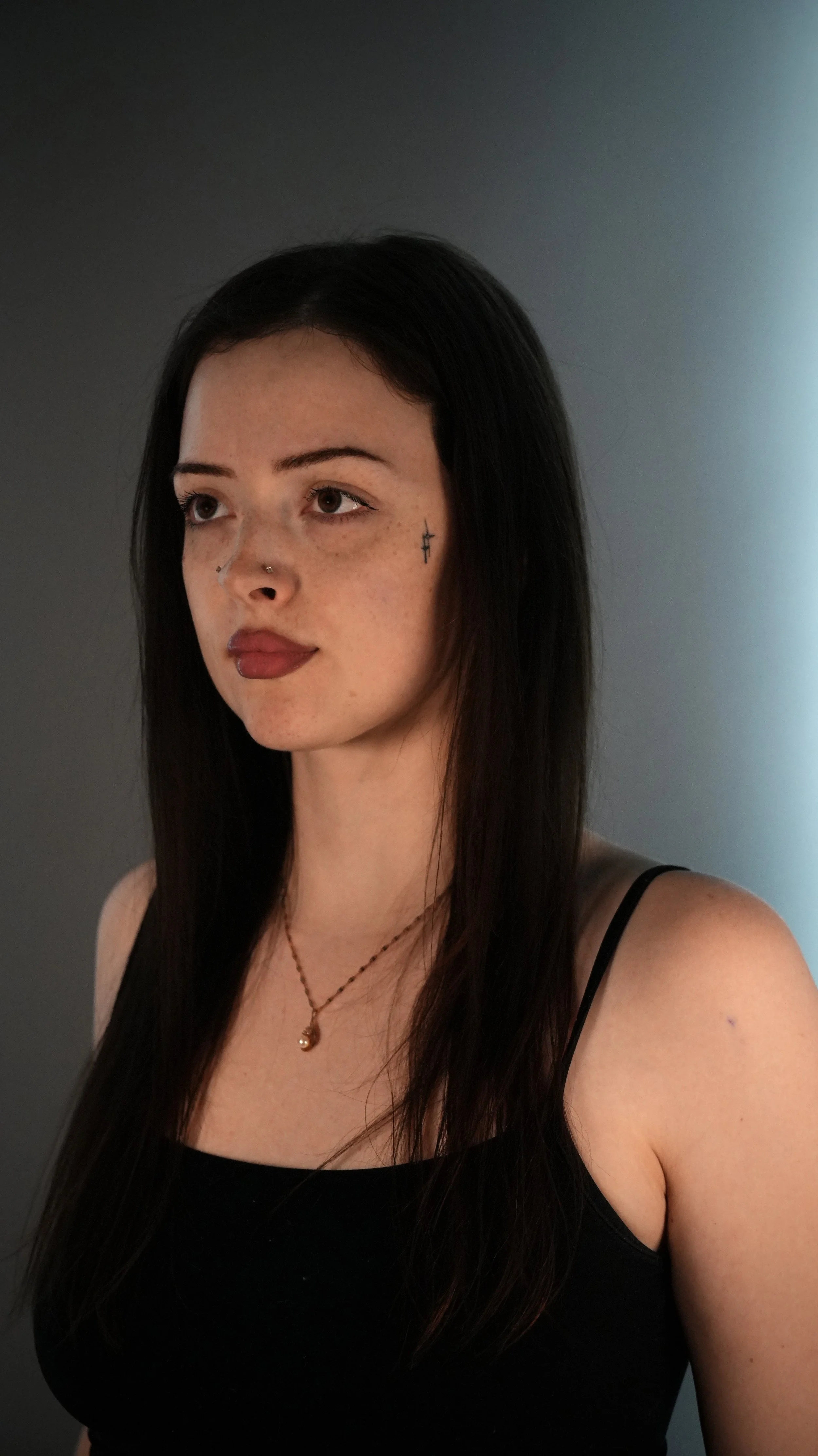 A young woman with long dark hair and facial tattoos, wearing a black tank top and a gold necklace with a crescent moon.