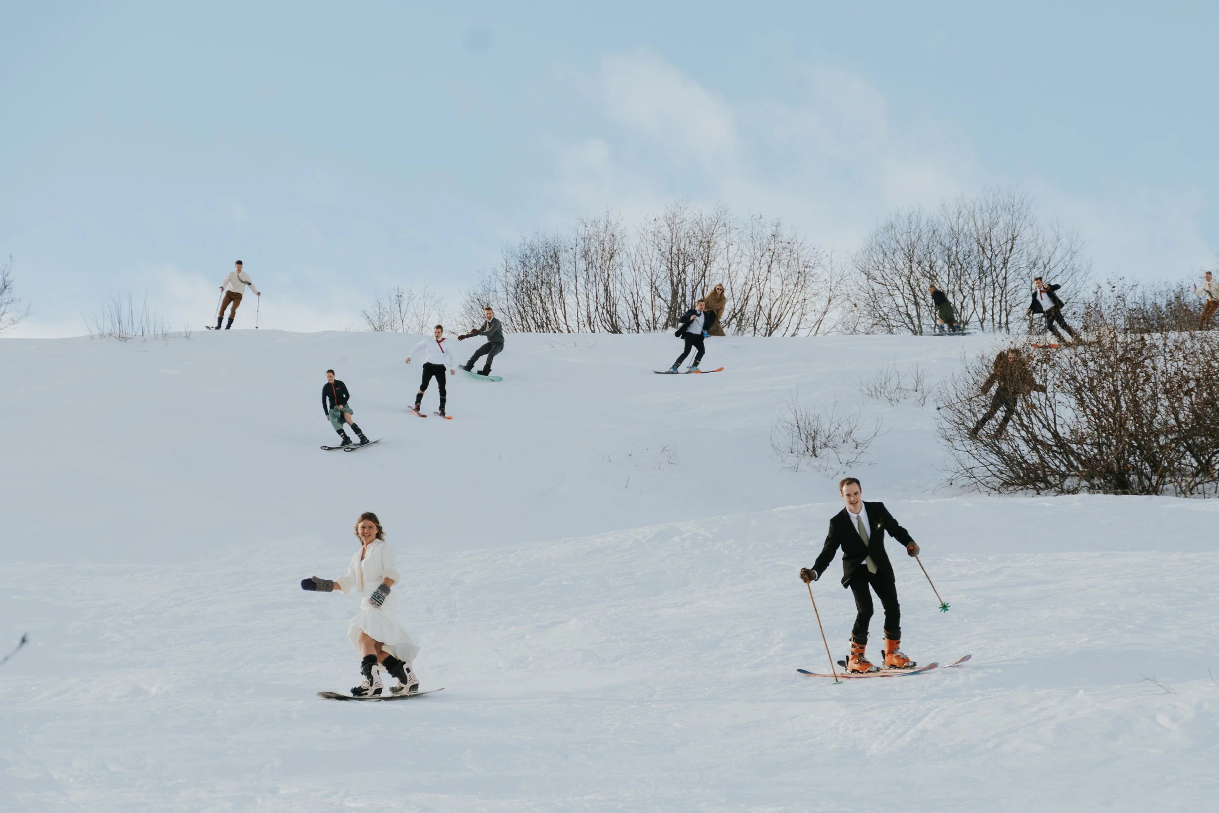 bride and groom in winter setting skiing and snowboarding down mountain with guests