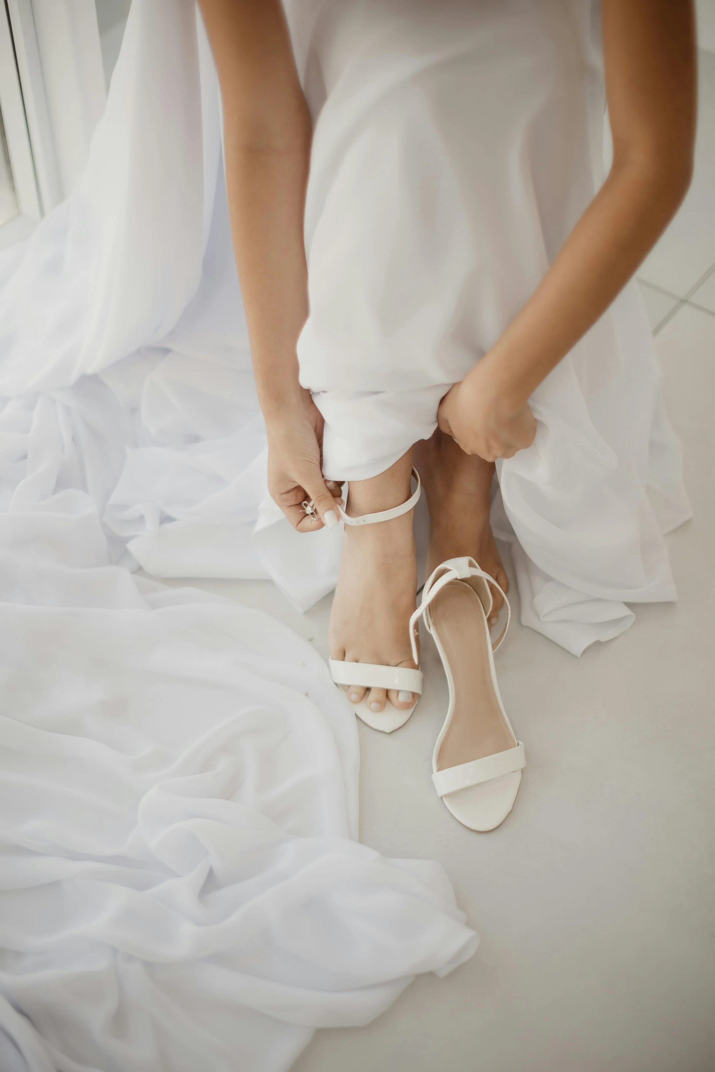 A person in a white dress is putting on white high-heeled sandals, with another pair of shoes on the floor nearby, on a light-colored floor with some draped white fabric.