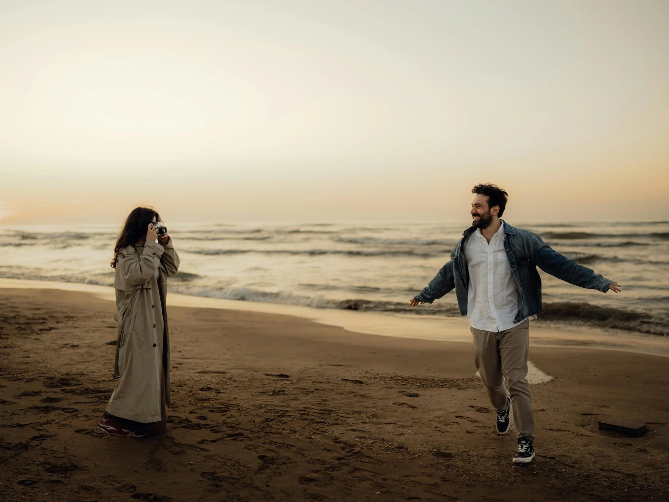 A woman in a beige trench coat taking a photo of a man in a denim jacket with a white shirt on the beach at sunset.