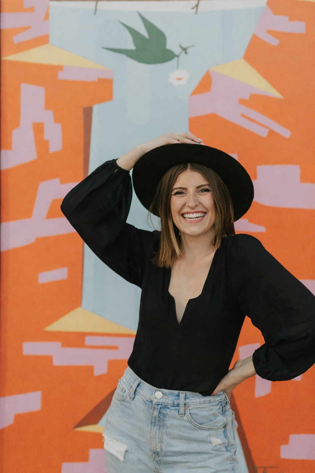 girl (mazzy)  holding hat in front of colorful mural