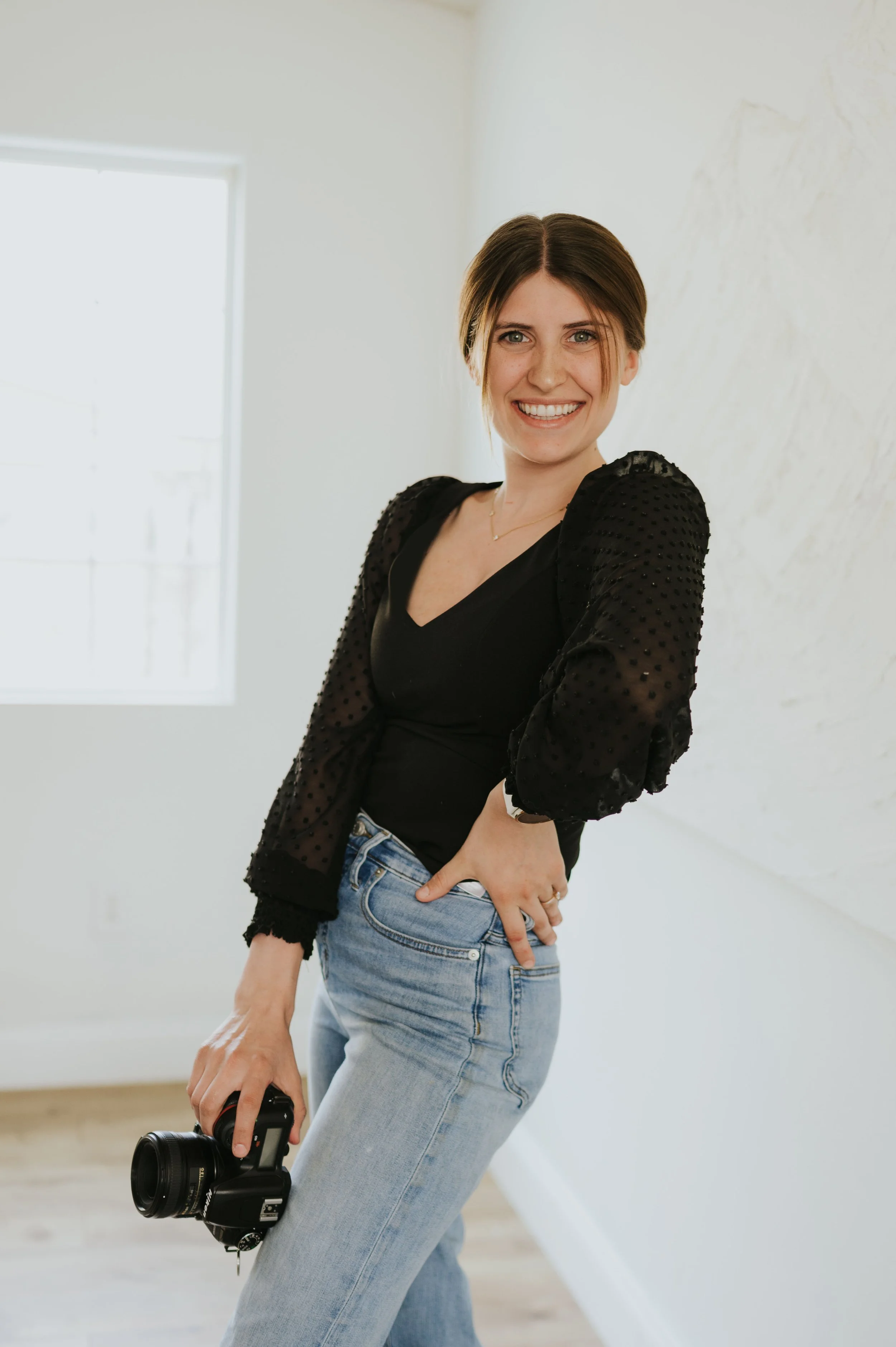A woman with brown hair in a casual black top and light blue jeans holds a camera while smiling indoors near a window and white wall.