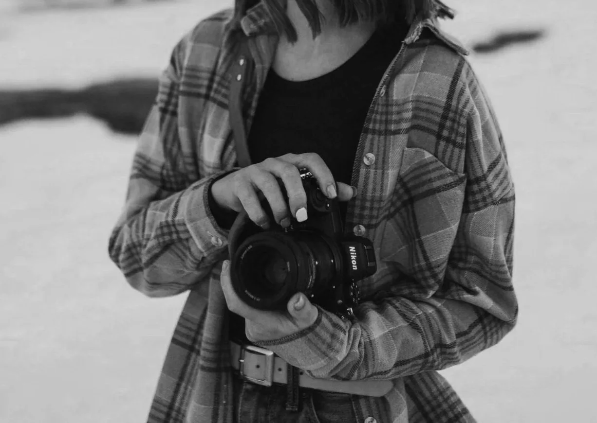 A woman holding a Nikon camera in a snowy outdoor setting, wearing a plaid jacket.