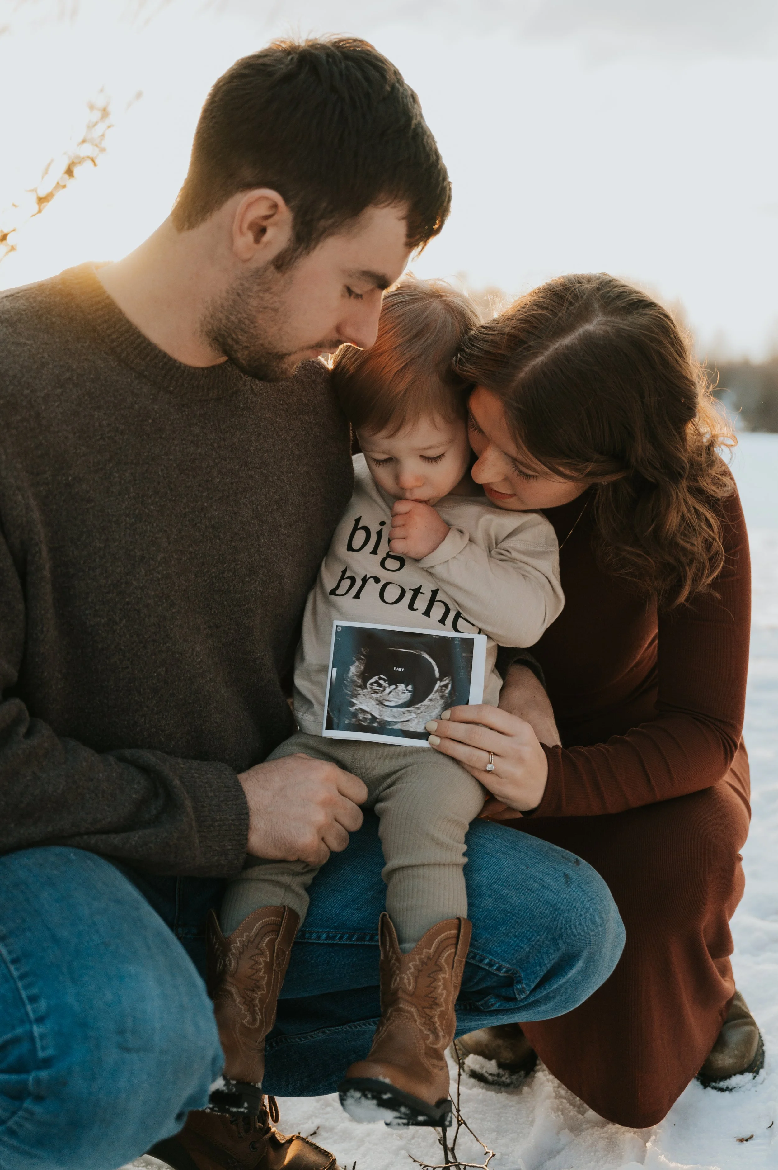 A family of three, a man, a woman, and a child, are sitting outside in the snow during sunset, sharing a moment of joy while holding an ultrasound photo of a baby.