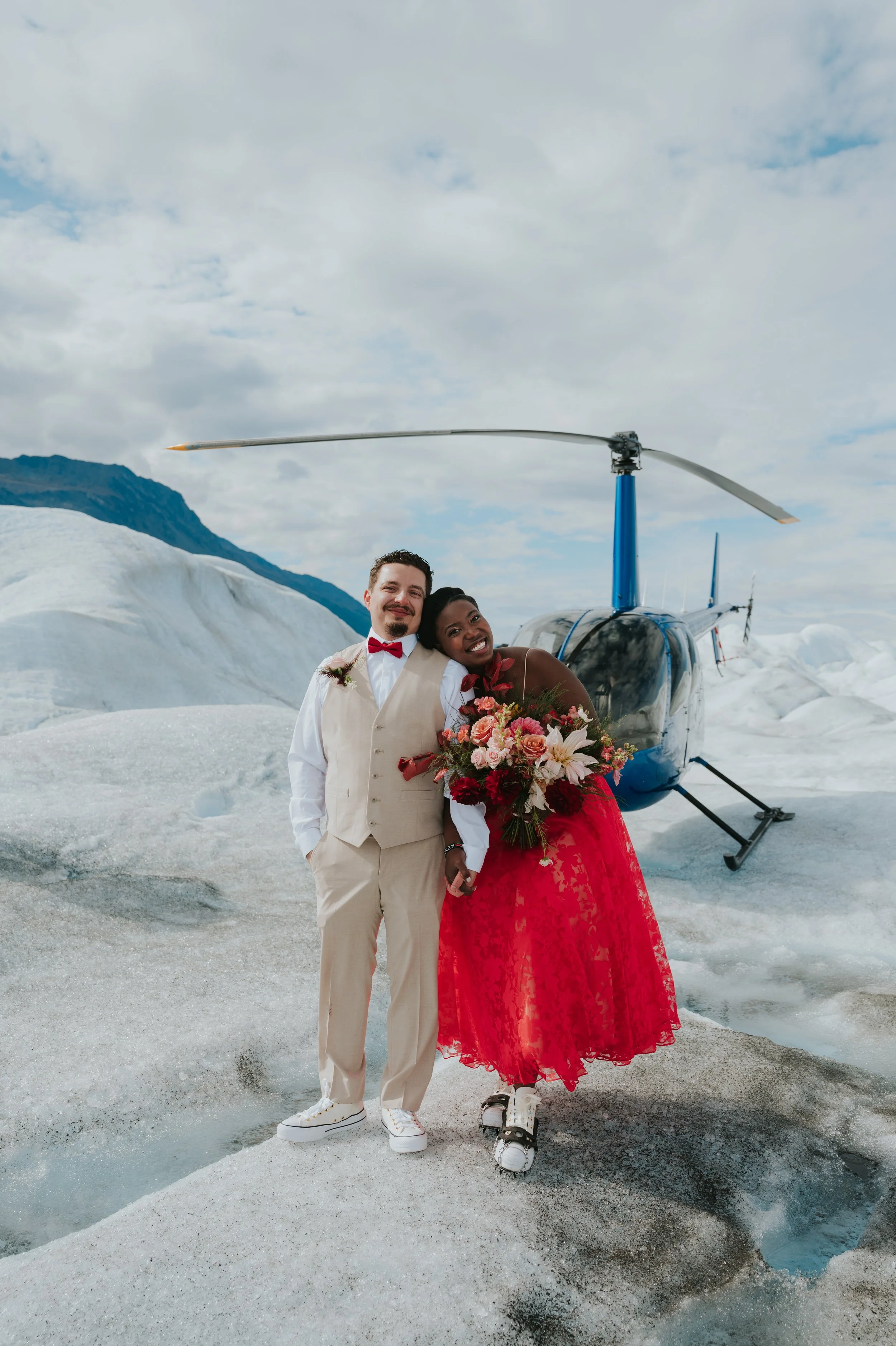 A couple in wedding attire, with the woman wearing ice skates, standing on ice near a helicopter in a glacier landscape with mountains and cloudy sky.