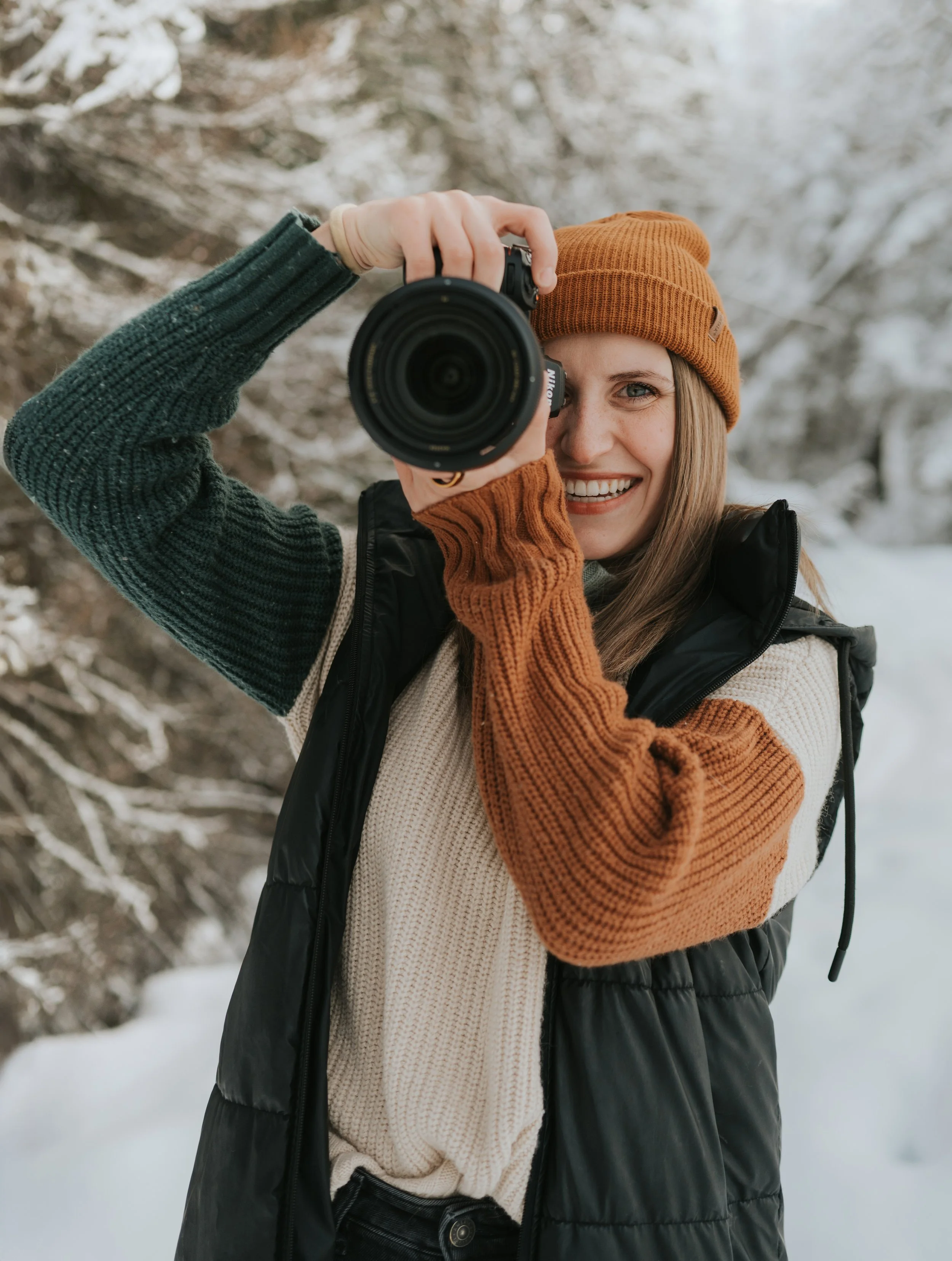 Photo of young woman photographer holding camera and smiling while wearing beanie and sweater in the winter outdoors