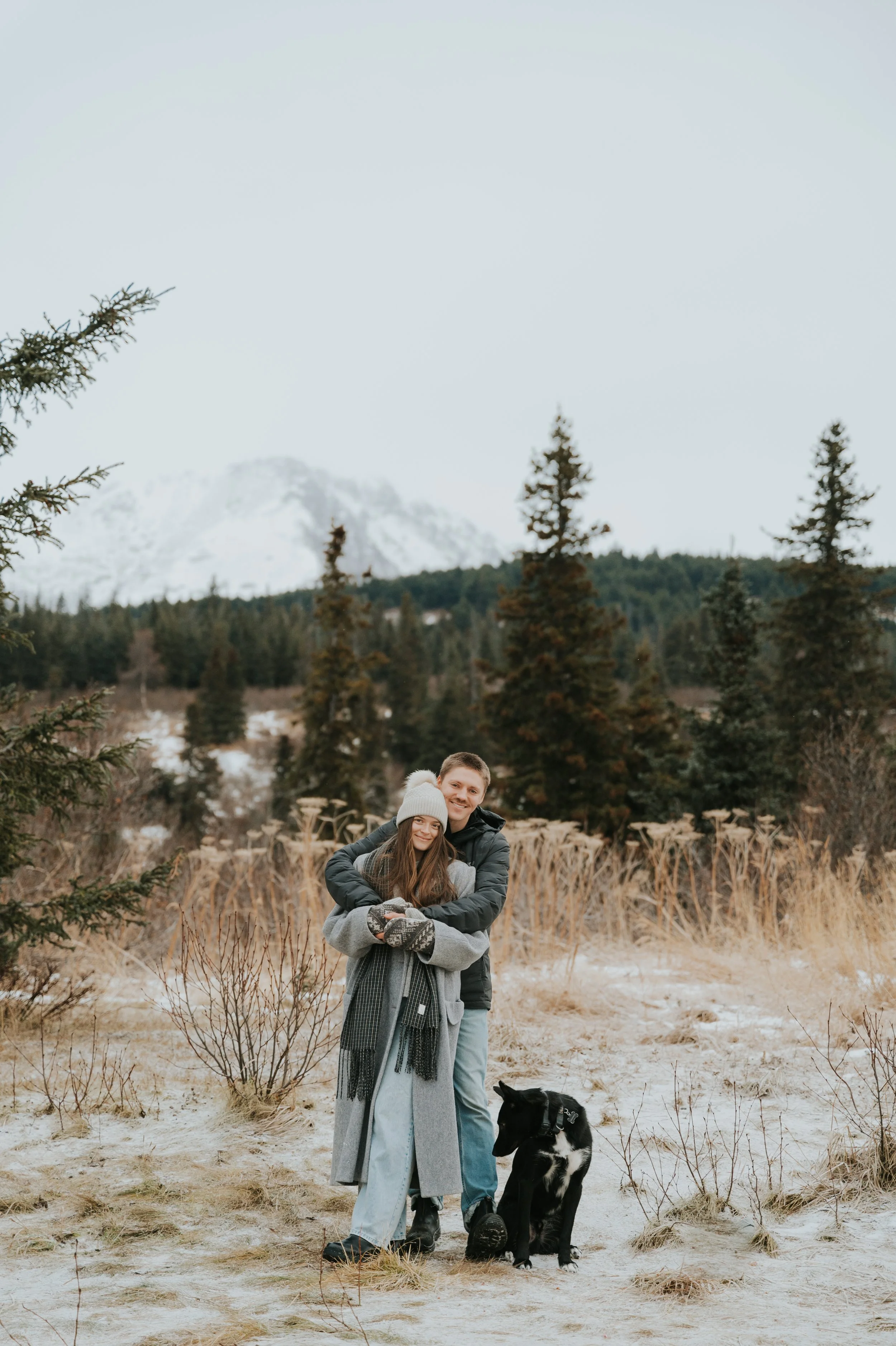 Cozy couple in winter setting smiling with dog at camera