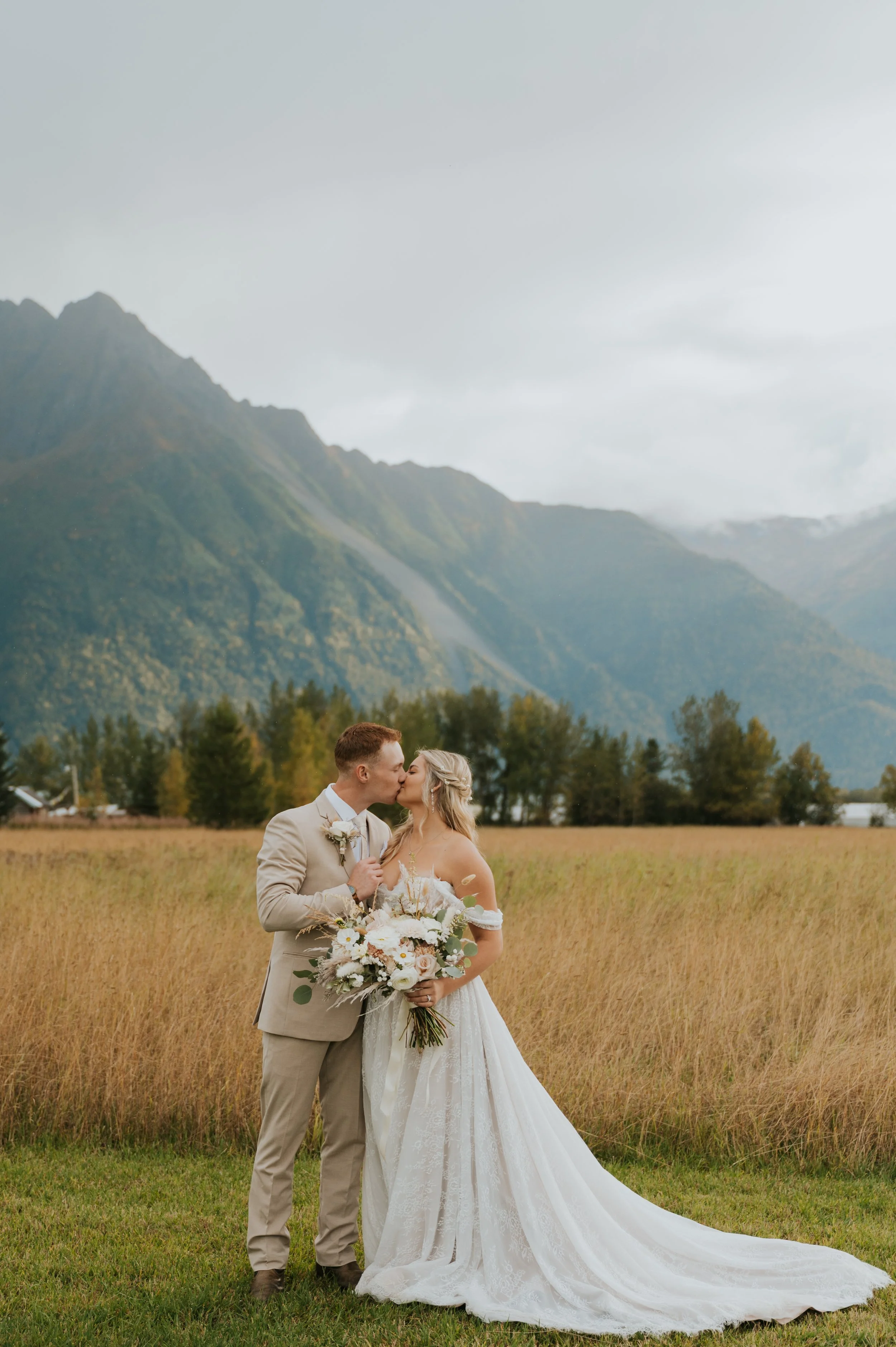 couple in wedding attire kissing in front of field and mountains