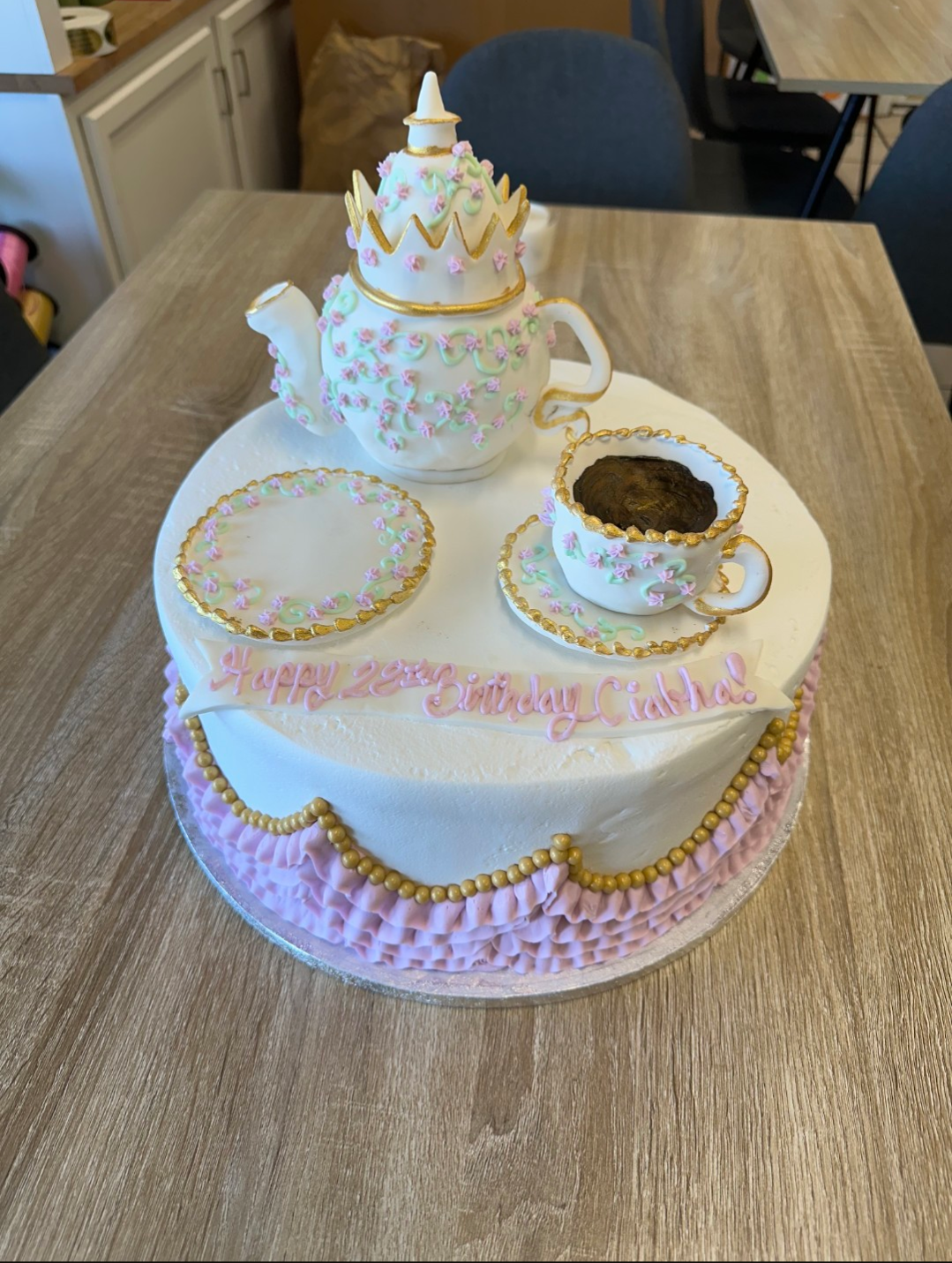A round birthday cake decorated as a tea set with a teapot, teacup, and saucer on top, all fondant covered with pink, green, and gold accents. The cake has a message written in pink icing: "Happy 22nd Birthday Cadie!" and is decorated with pink ruffl
