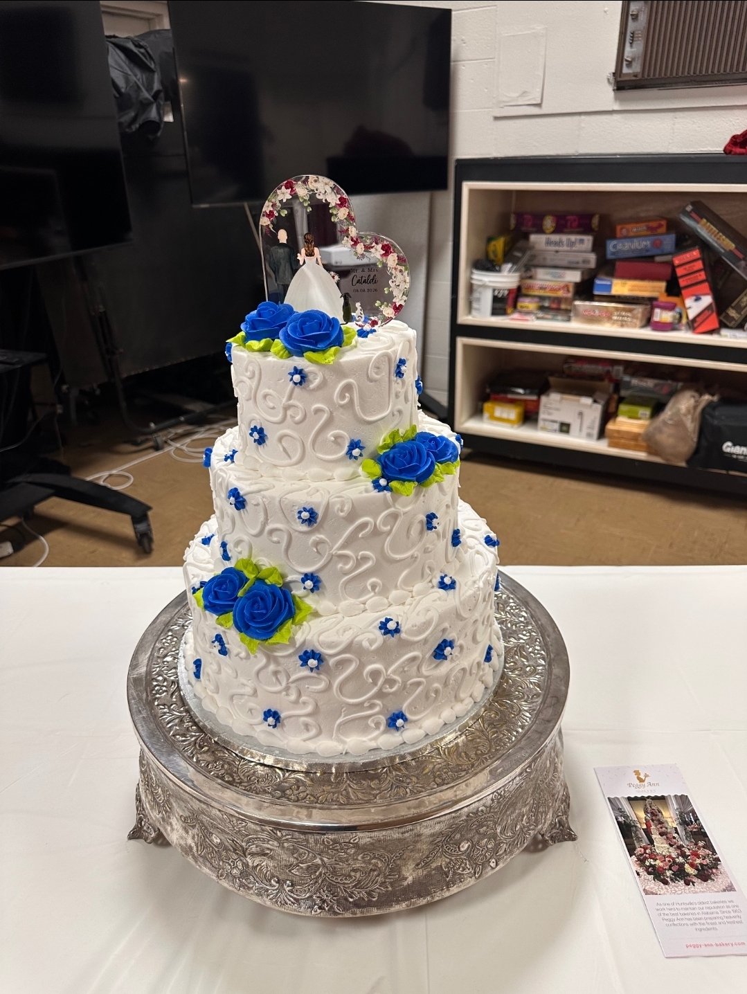 Three-tier white wedding cake with blue roses and small blue flowers, topped with a wedding photo cake topper, displayed on an ornate silver cake stand in a room with a white table and various shelves in the background.