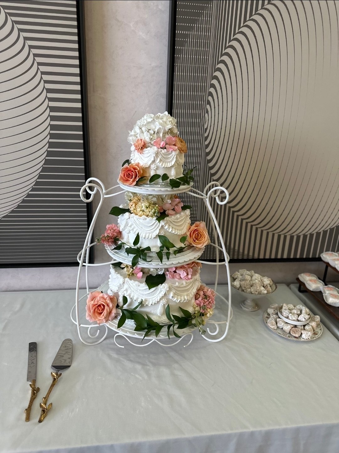 A three-tiered wedding cake decorated with white icing and pink and peach roses, green leaves, and other flowers, on a white metal stand in front of a modern patterned wall, with dessert tongs and plates of sweets on the table.