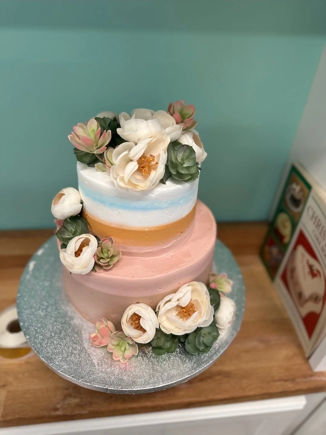 Two-tiered cake with pastel pink, yellow, and white frosting, decorated with artificial flowers and succulents on top and around the base.
