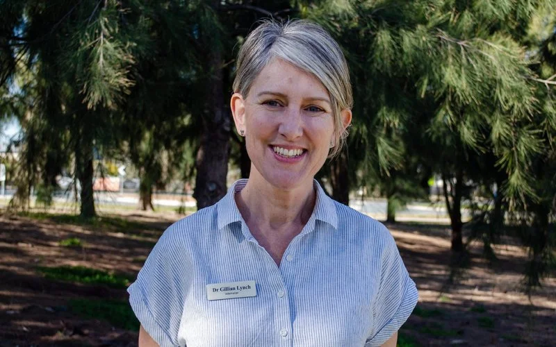 A smiling woman with short blonde hair standing outdoors in front of trees, wearing a light blue striped collared shirt with a name tag that reads "Dr. Gillian Lynch."