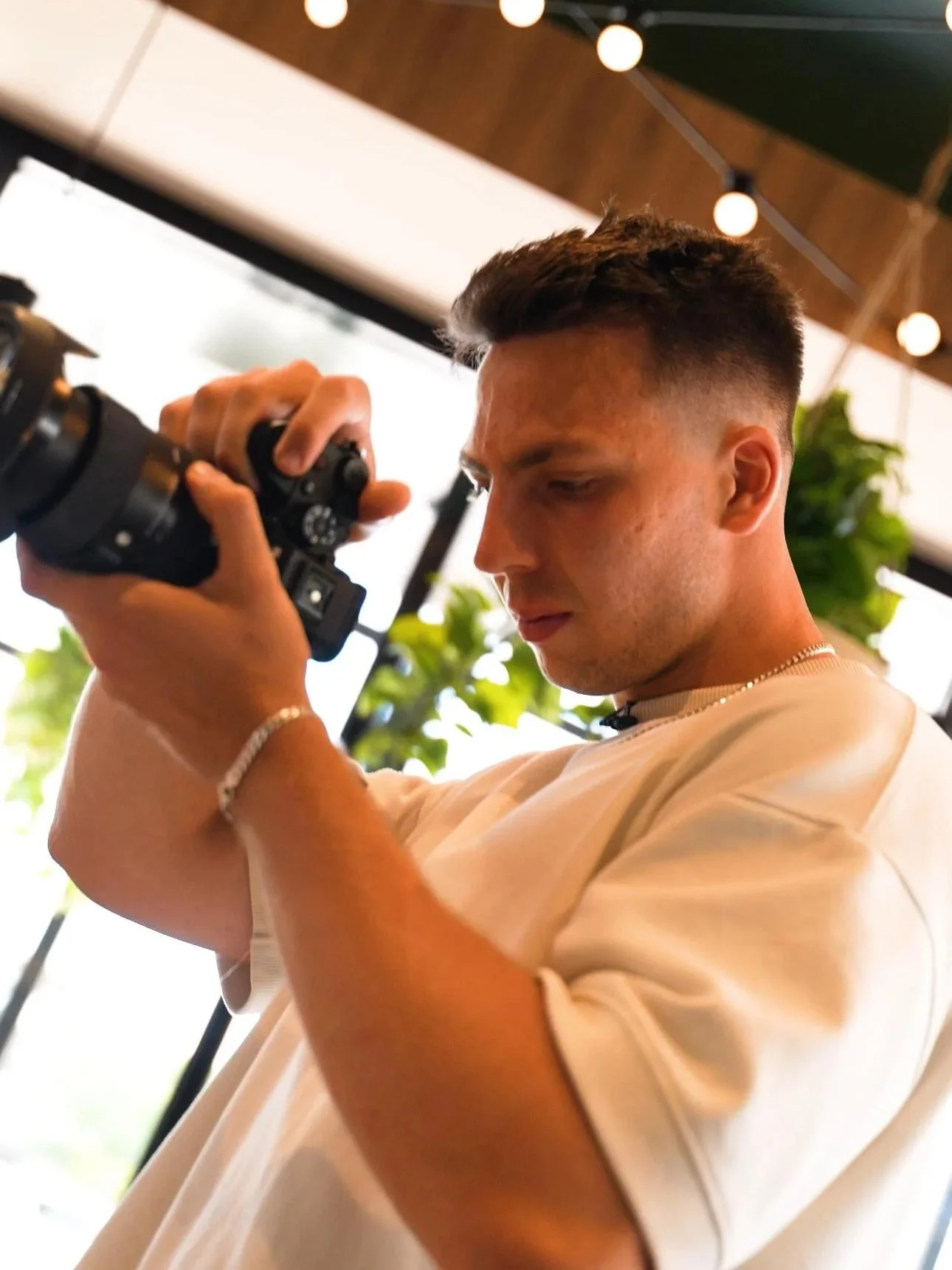 A young man with short dark hair is adjusting the settings on a black digital camera, looking down at it. He is wearing a light-colored T-shirt, a bracelet on his left wrist, and a necklace. The background shows indoor decor with hanging string light