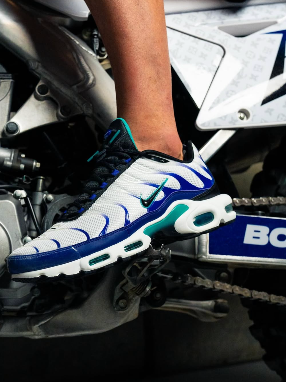 Close-up of a person's foot wearing a white and blue Nike sneaker resting on a motorcycle pedal.