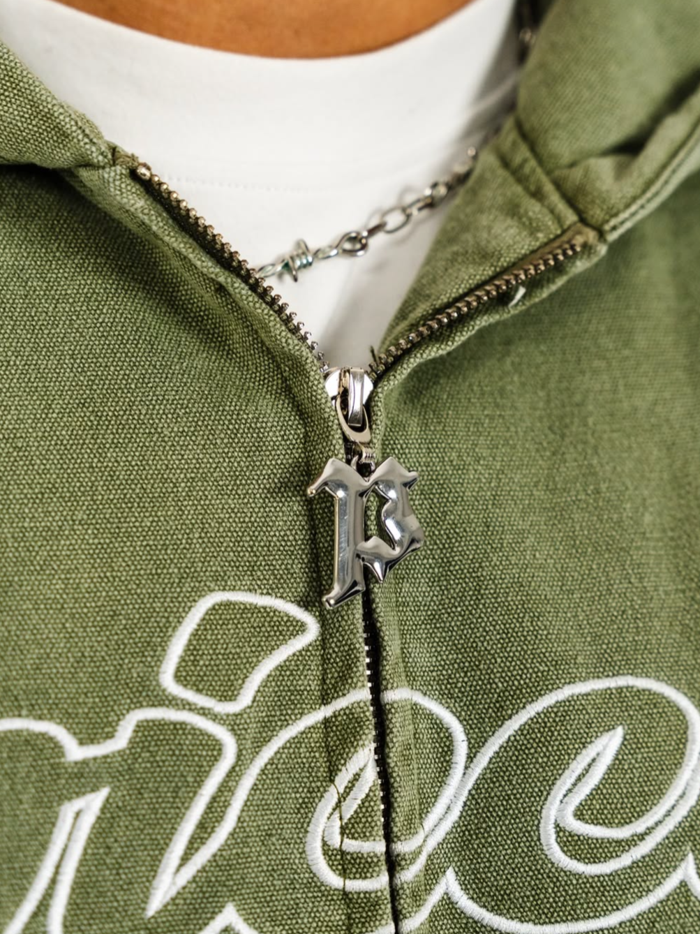 Close-up of a person wearing a green hoodie with white embroidery and a silver necklace with a pendant of the letter 'K' in a gothic font, partially unzipped to reveal a white shirt underneath.