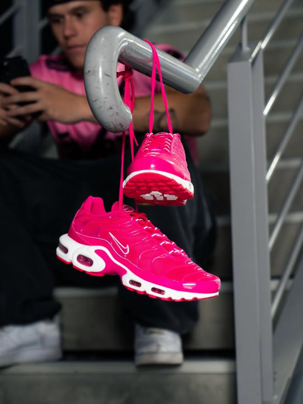 Pink Nike sneakers hanging from a stair railing with a person sitting in the background using a phone.