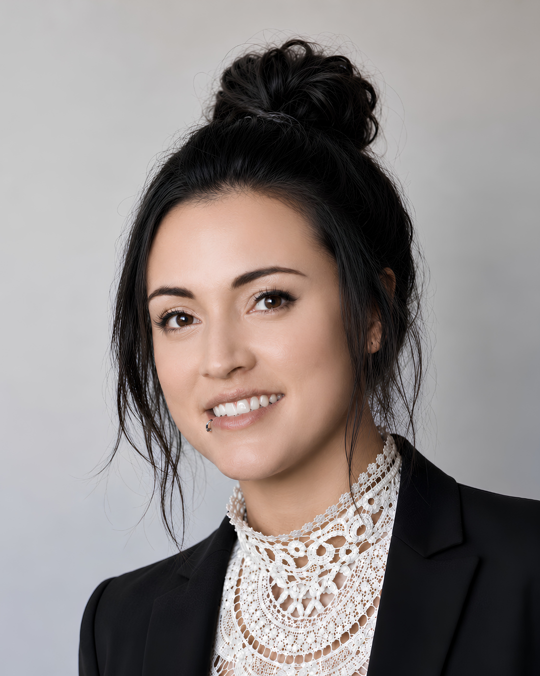Close-up portrait of a woman with dark hair styled in a messy bun, smiling, wearing a black blazer over a white lace top, against a plain light grey background.