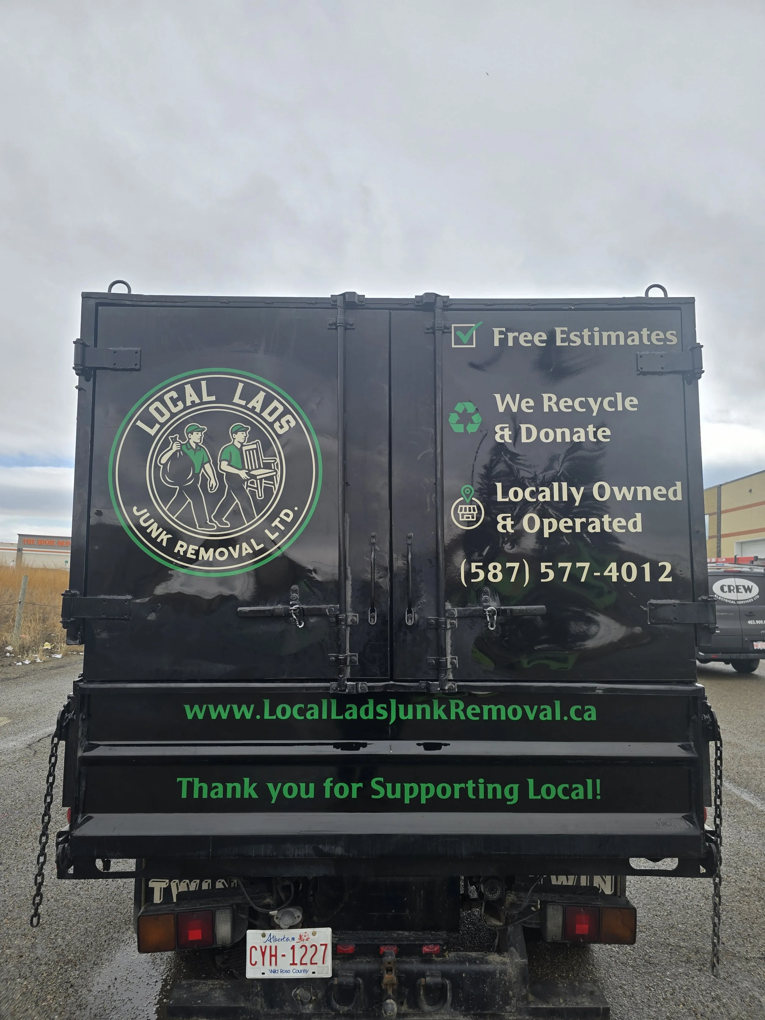Black junk removal truck with green and white lettering and logo, advertising Local Lads Junk Removal Ltd. with services including free estimates, recycling, donation, locally owned and operated, and contact information. The website is www.LocalLadsJunkRemoval.ca and the slogan 'Thank you for Supporting Local!' is displayed.