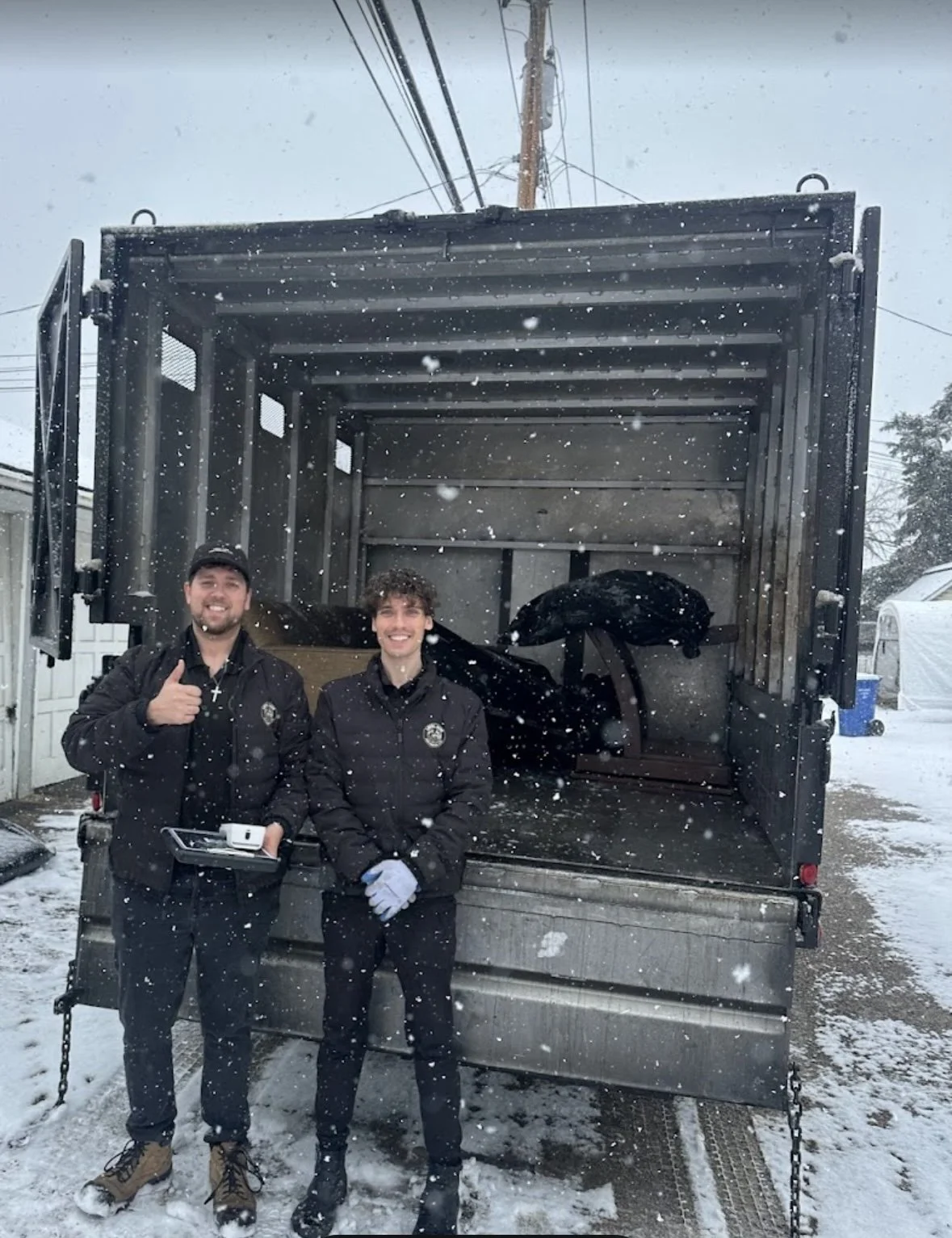 Two men in black jackets standing in front of an open delivery truck on a snowy day, with snow falling and snow on the ground.