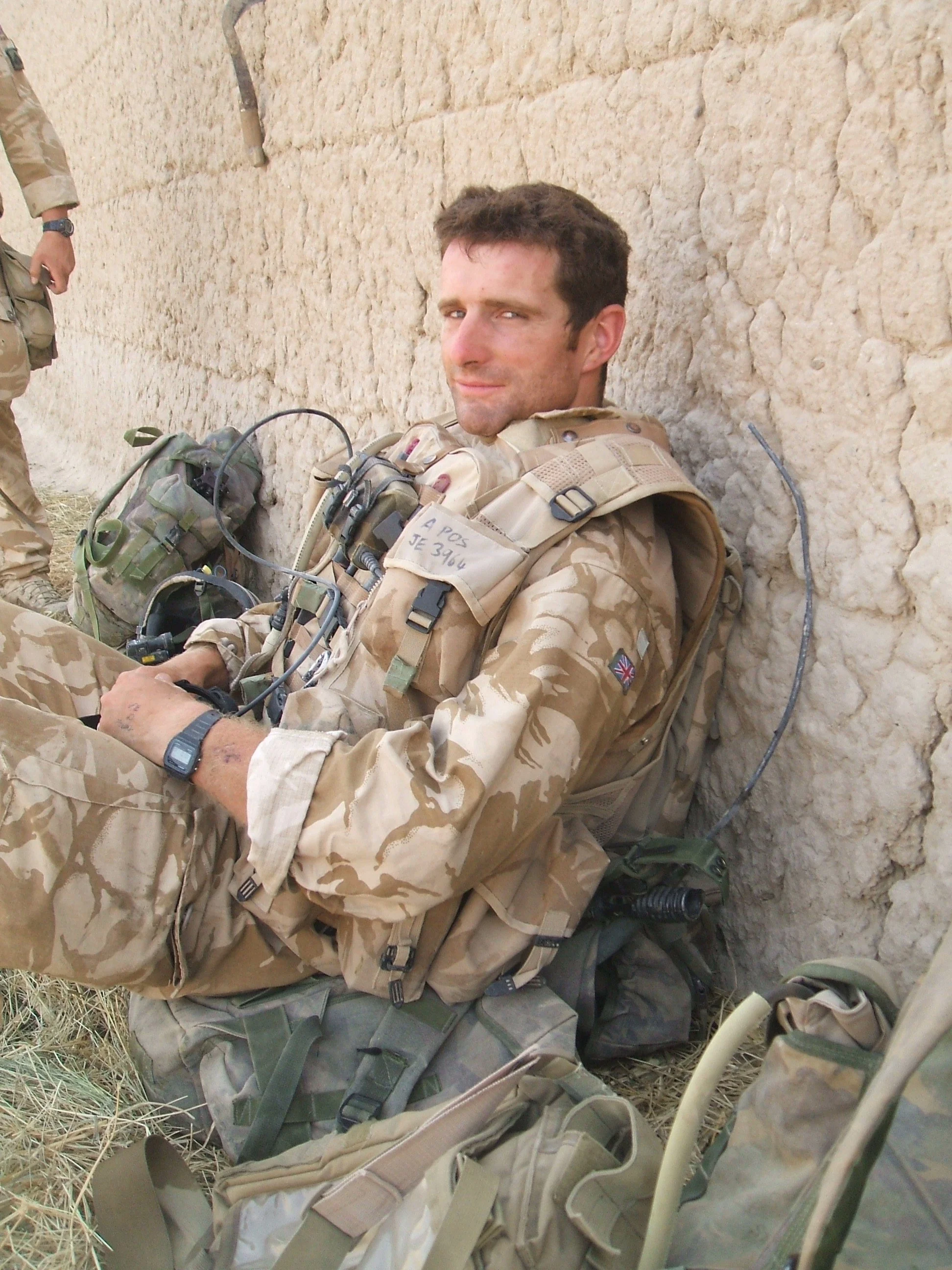 A soldier sitting against a wall with desert camouflage uniform, equipment, and tactical gear, looking at the camera.
