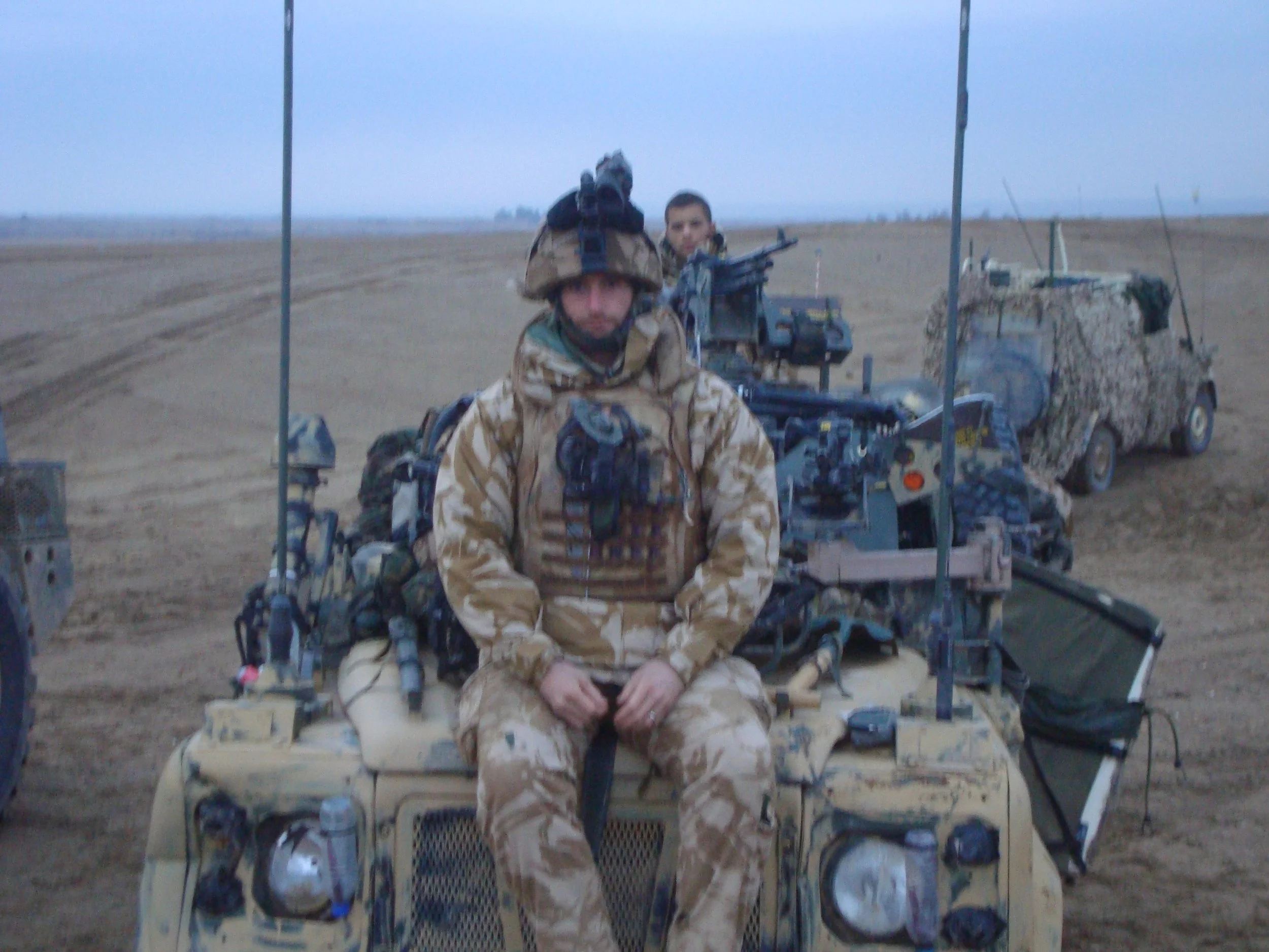 Soldier in camouflage sitting on military vehicle in desert environment with another person in the background.