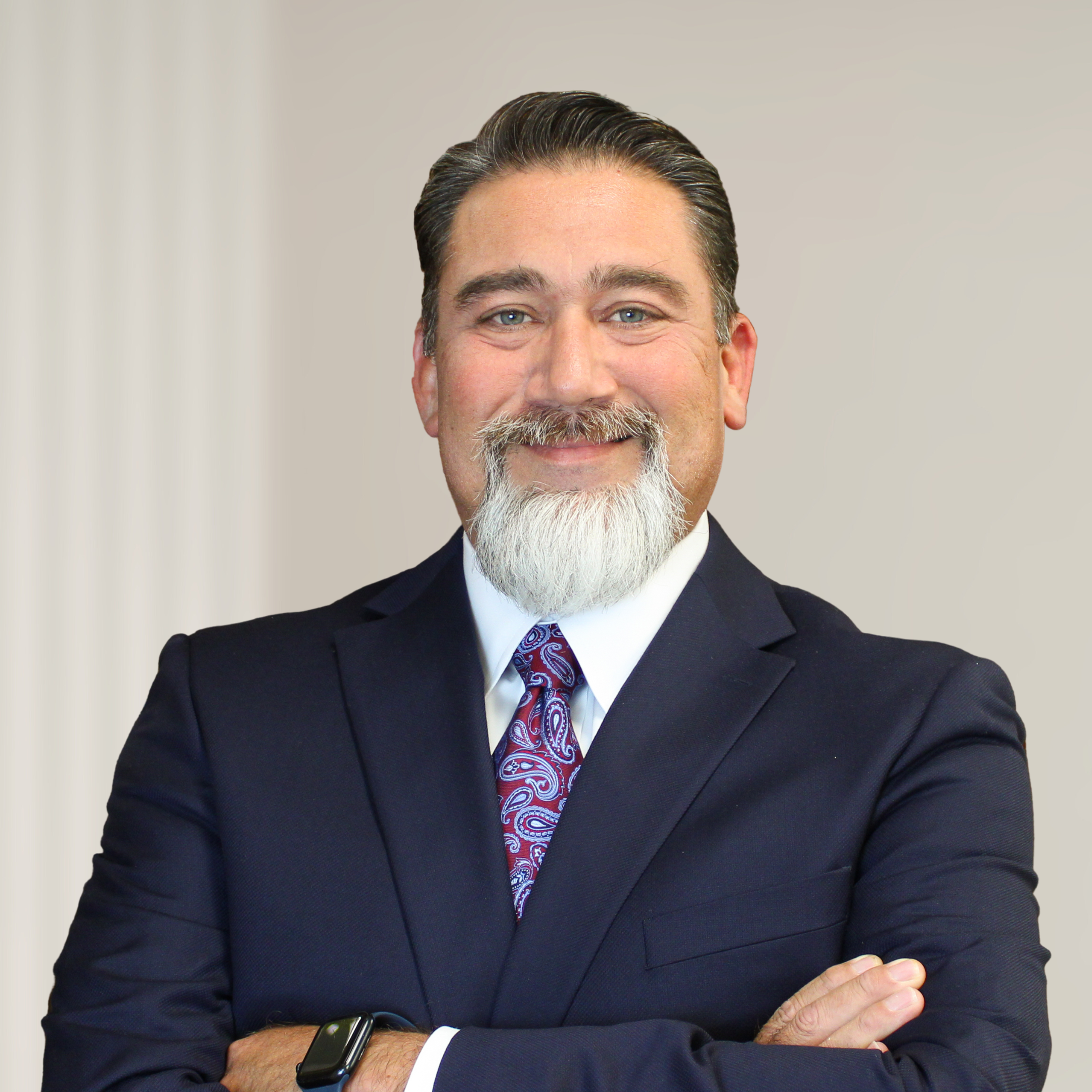 A middle-aged man with a beard and mustache wearing a dark suit, white shirt, and a patterned tie standing with arms crossed and smiling.