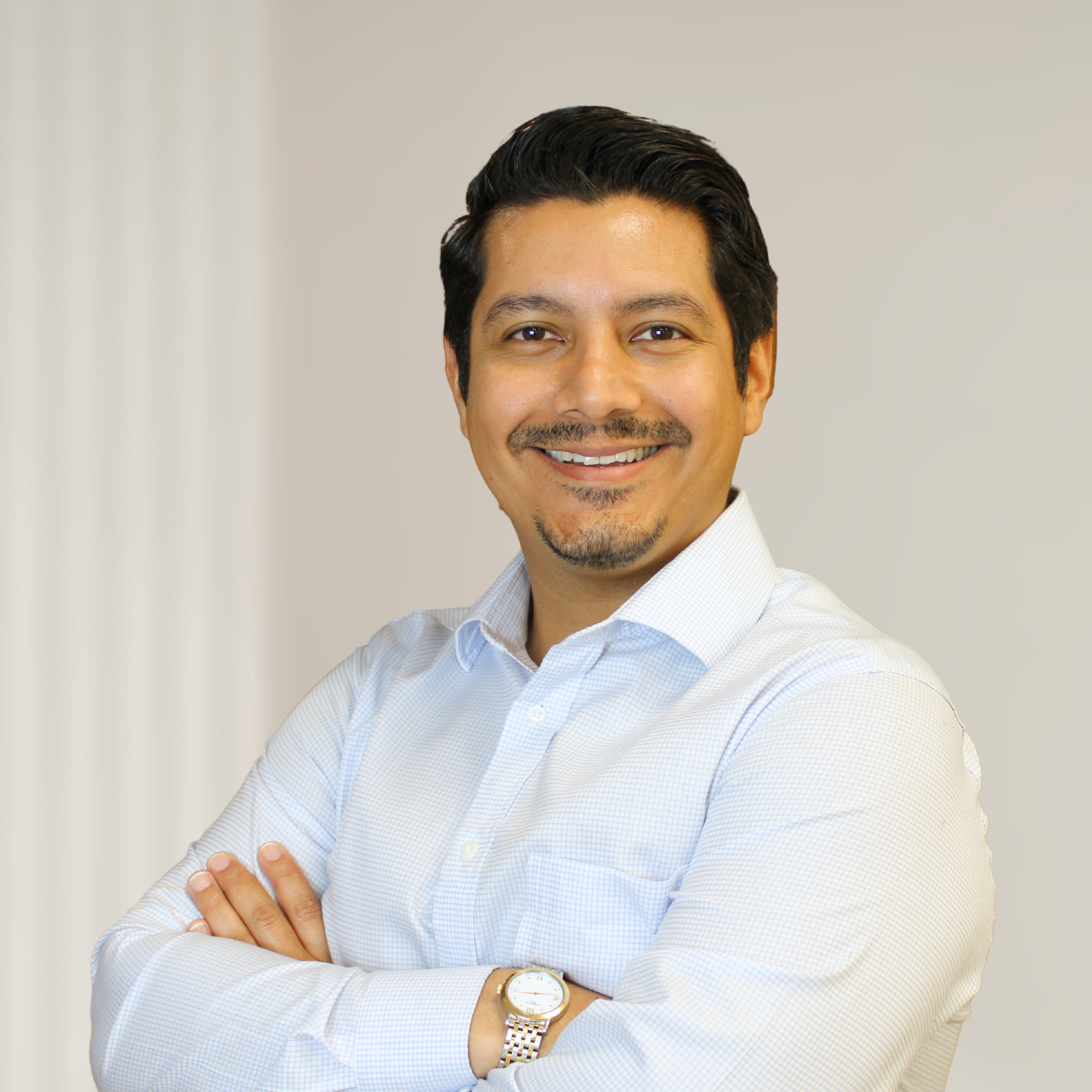 Portrait of a smiling man with dark hair, wearing a white dress shirt, crossed arms, standing in front of a wooden background.
