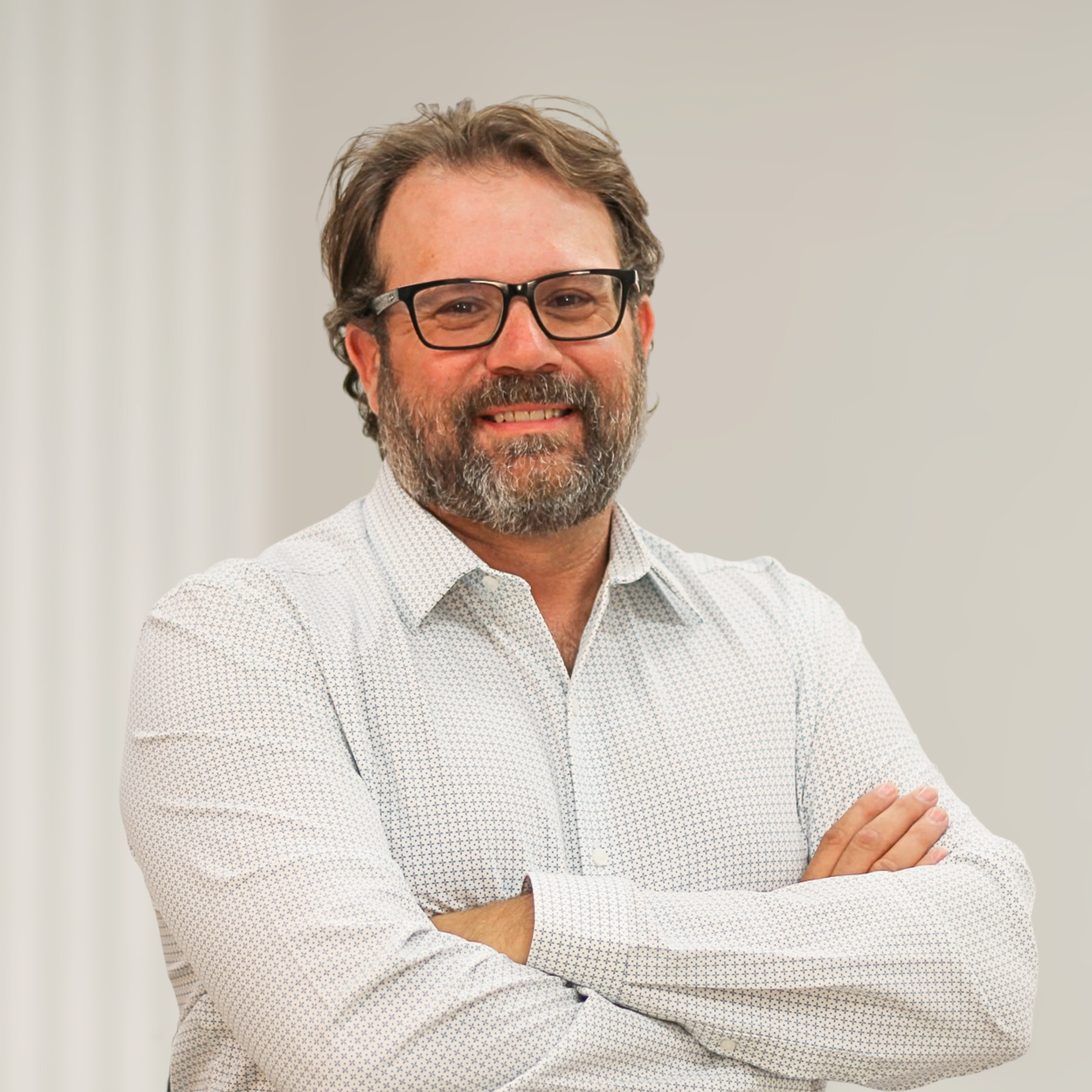 Smiling man with glasses and a beard, wearing a white shirt, standing with arms crossed in front of a wooden panel background.