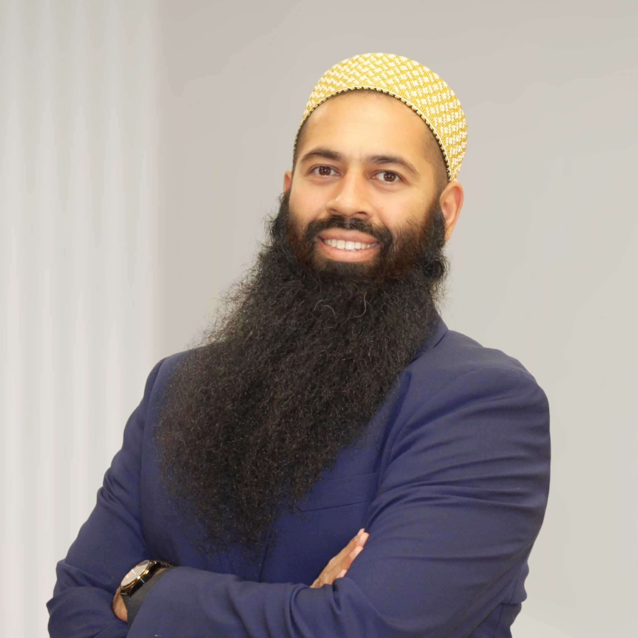 A man with a full, beard and mustache wearing a traditional white and gold cap and a grey suit, standing in front of a wooden background.