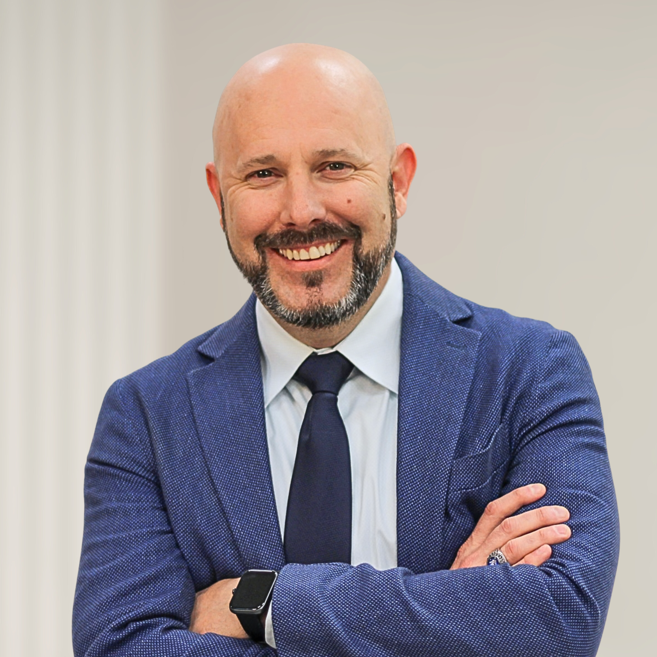 A man with a bald head and beard wearing a black suit with a white shirt standing with arms crossed in front of a wooden background.
