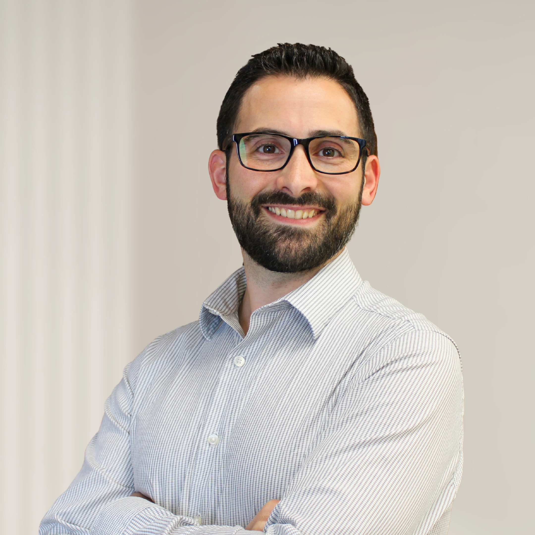 A smiling man with dark hair, a beard, and glasses, wearing a light-colored, checked dress shirt, standing with his arms crossed in front of a wooden decorative background.