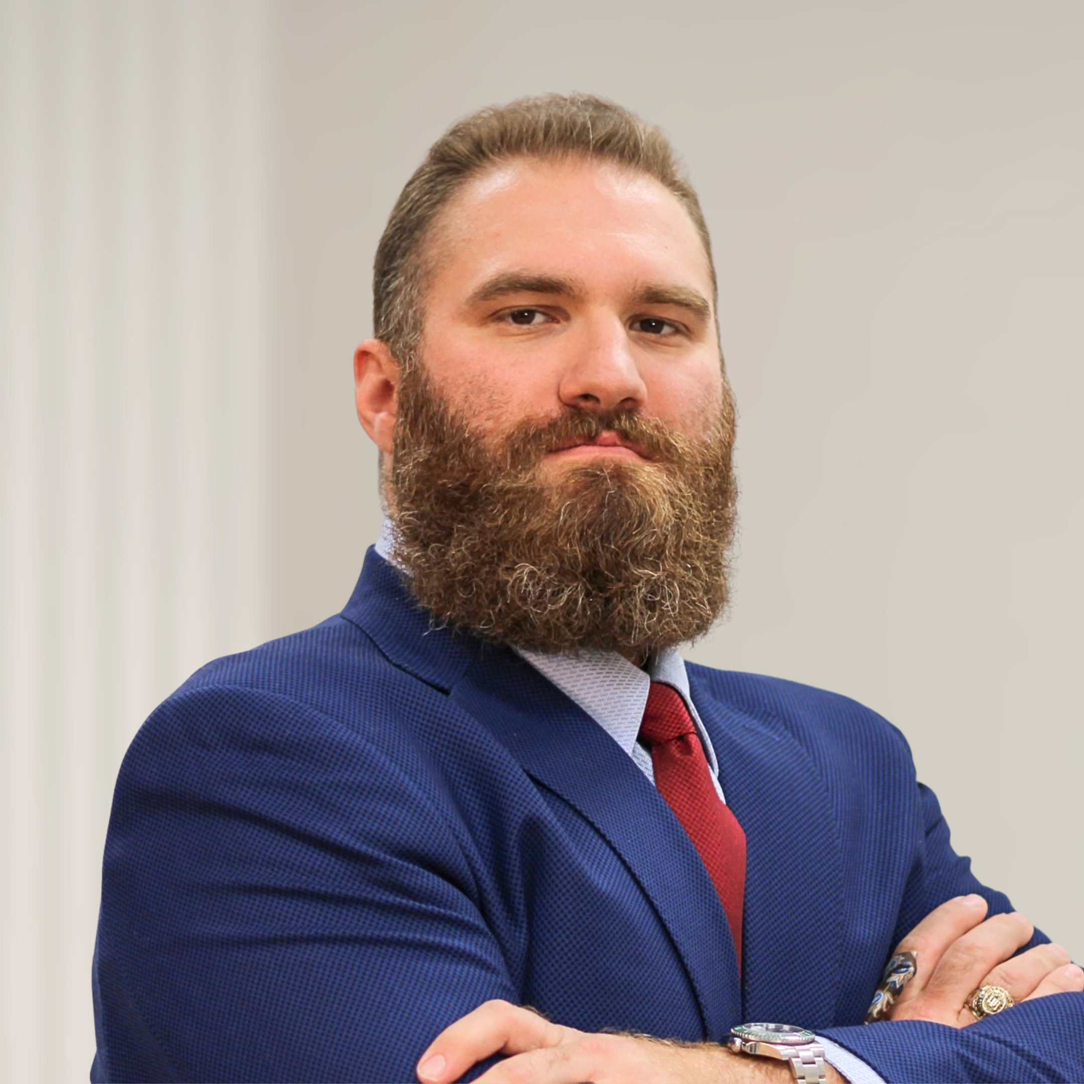 A man with a well-groomed beard and slicked-back hair wearing a dark blue suit, white shirt, and purple patterned tie, standing in front of a wooden background.