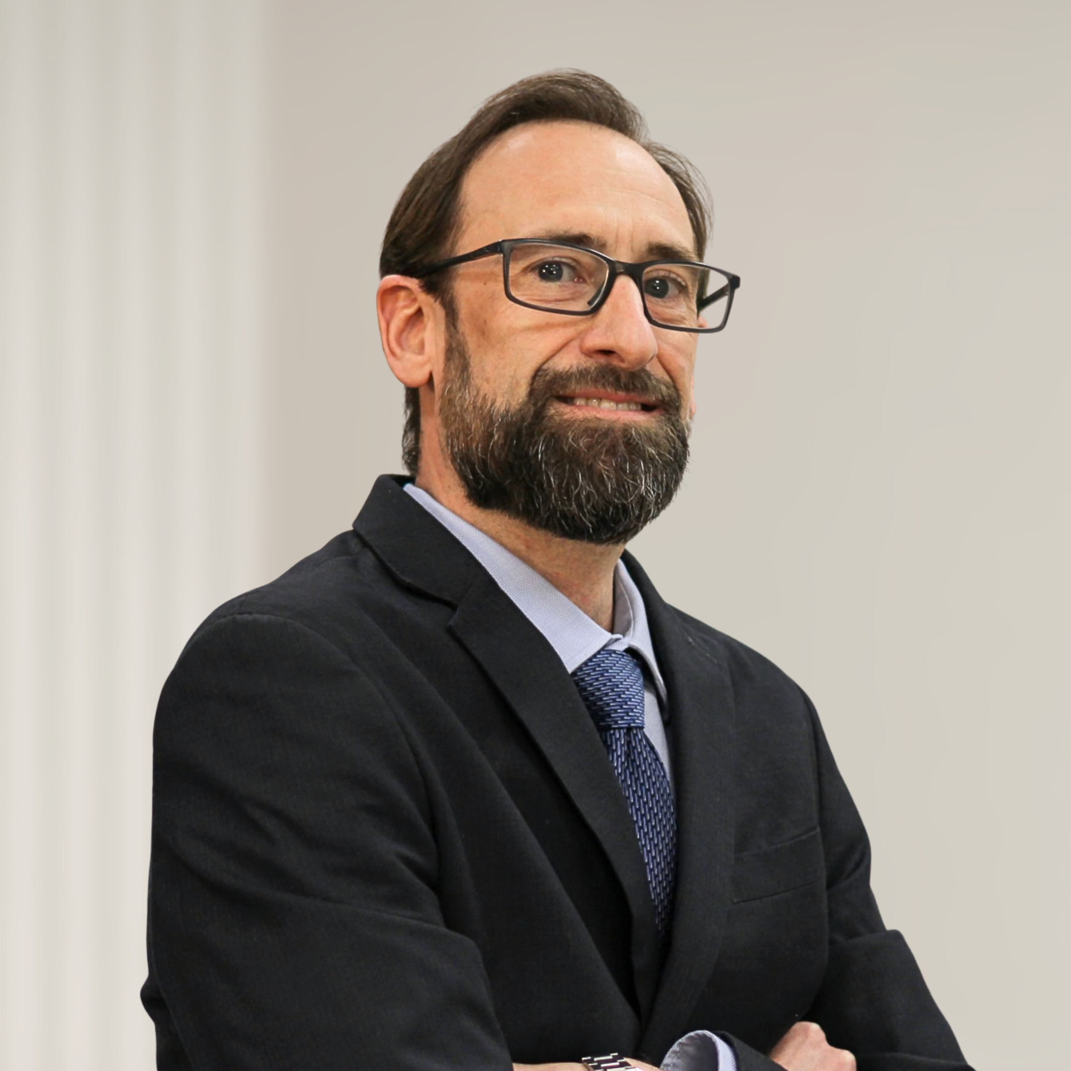 A man with a beard and short brown hair wearing a dark suit, light blue shirt, and patterned tie, standing in front of a wooden background.