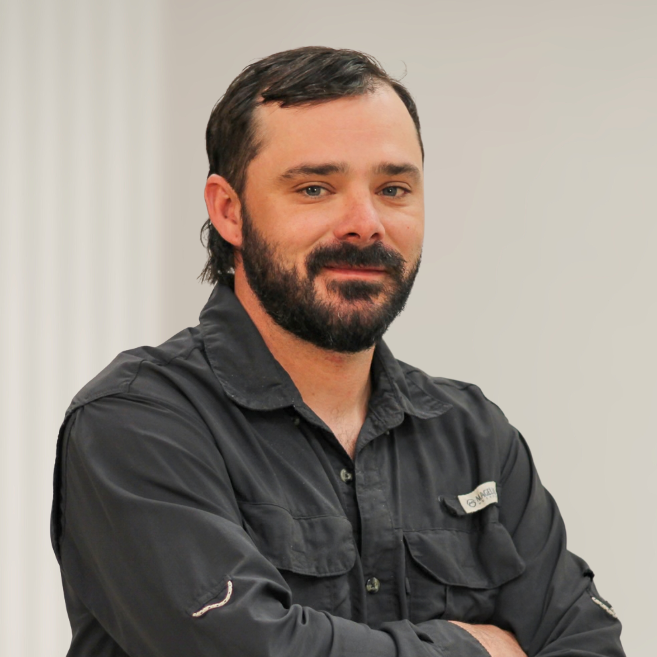 A man with dark hair and a beard, wearing a black vest over a plaid shirt, standing with arms crossed in front of a wooden background.