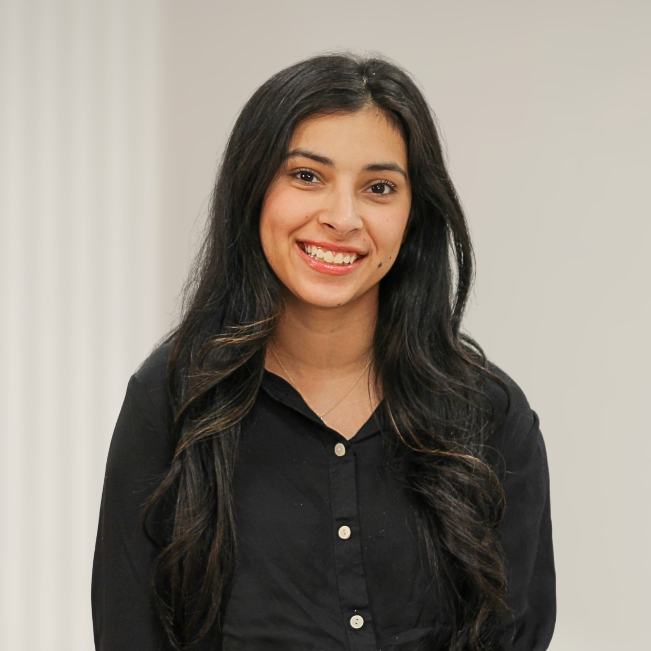 A woman with long wavy black hair, wearing a white striped button-up shirt, smiling and crossing her arms, standing in front of a wooden background.