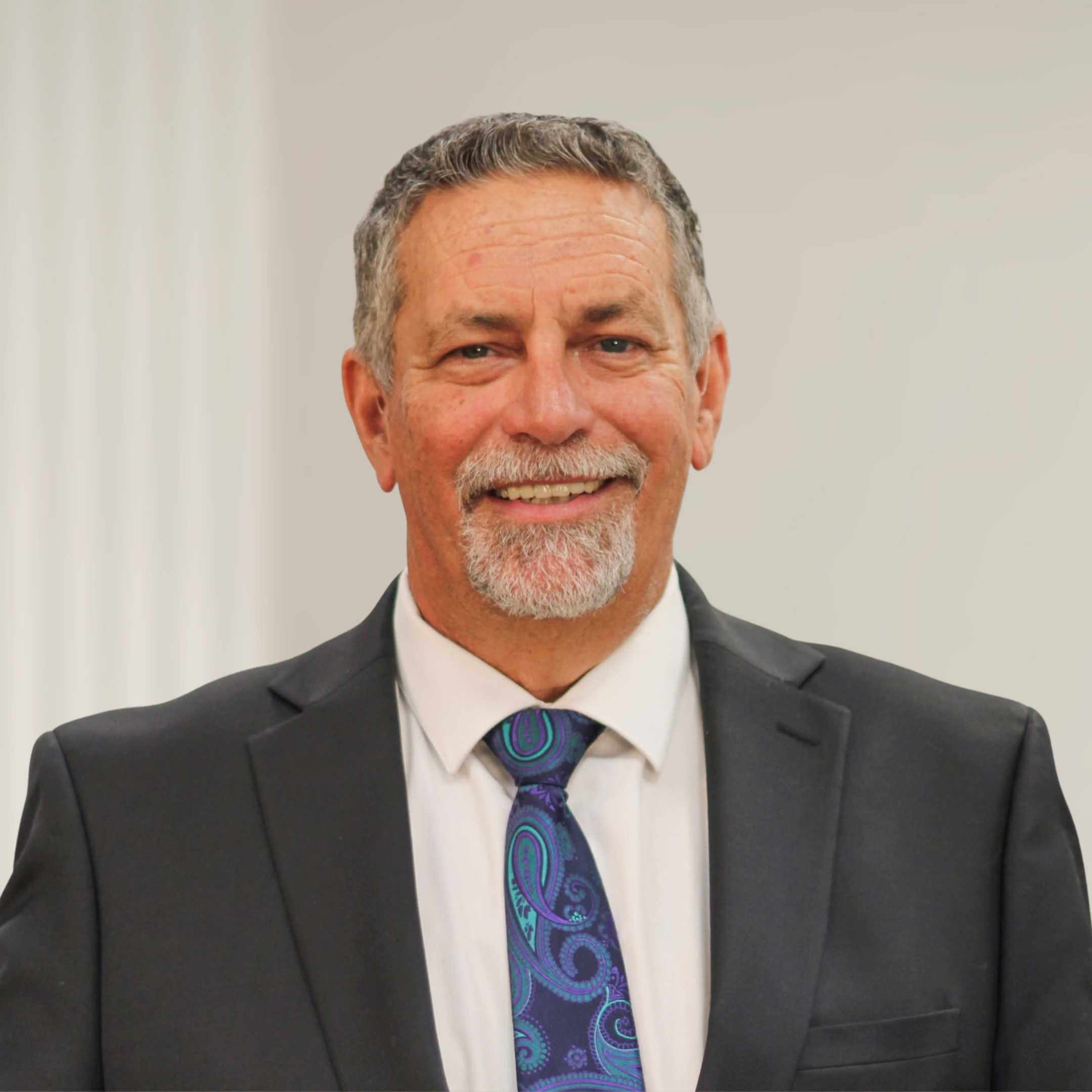 A smiling middle-aged man with gray hair and a beard, dressed in a dark suit, white shirt, and patterned tie, standing in front of a wooden background.