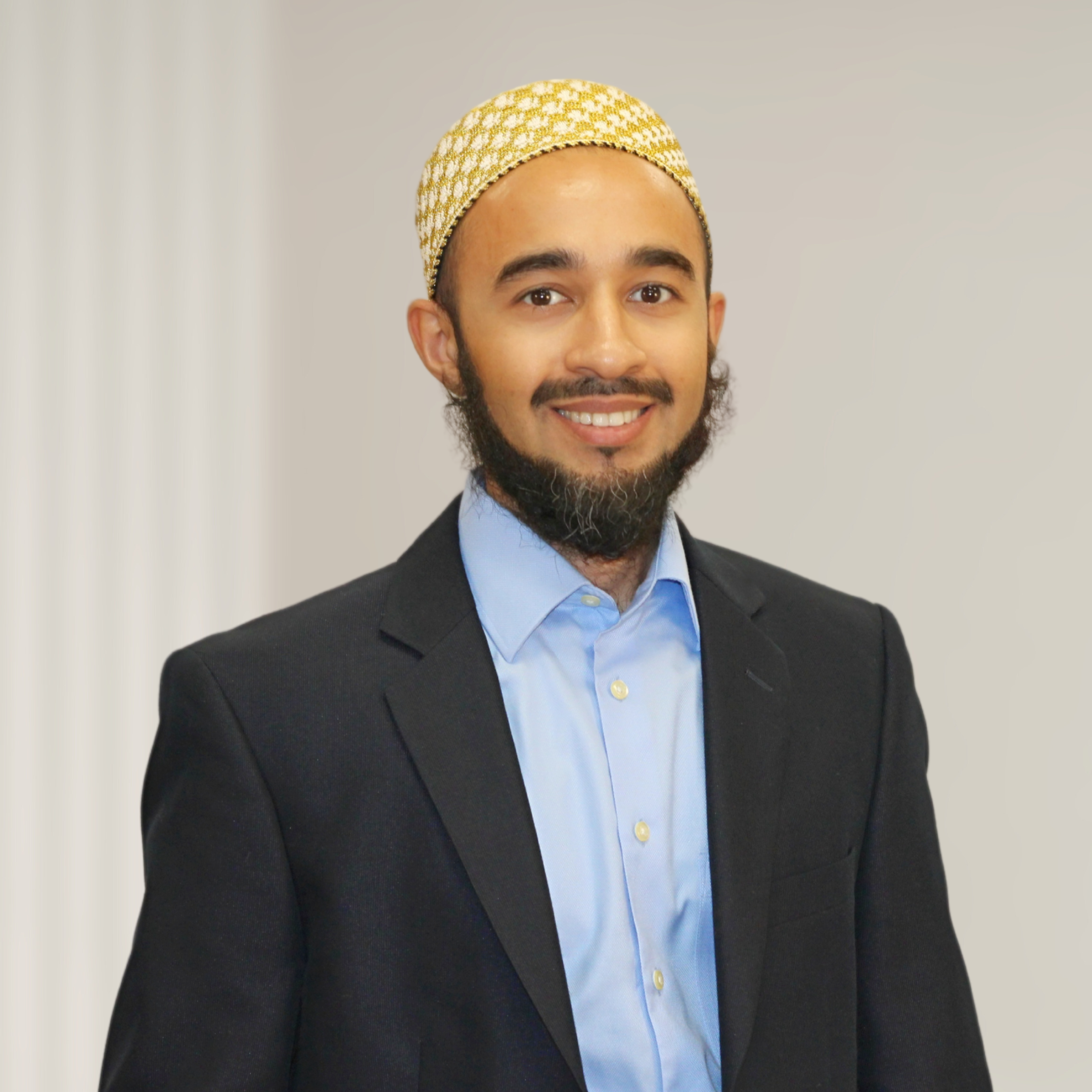 A man with a beard and mustache wearing a blue dress shirt, black blazer, and a white kufi hat, standing in front of a wooden wall with vertical slats.